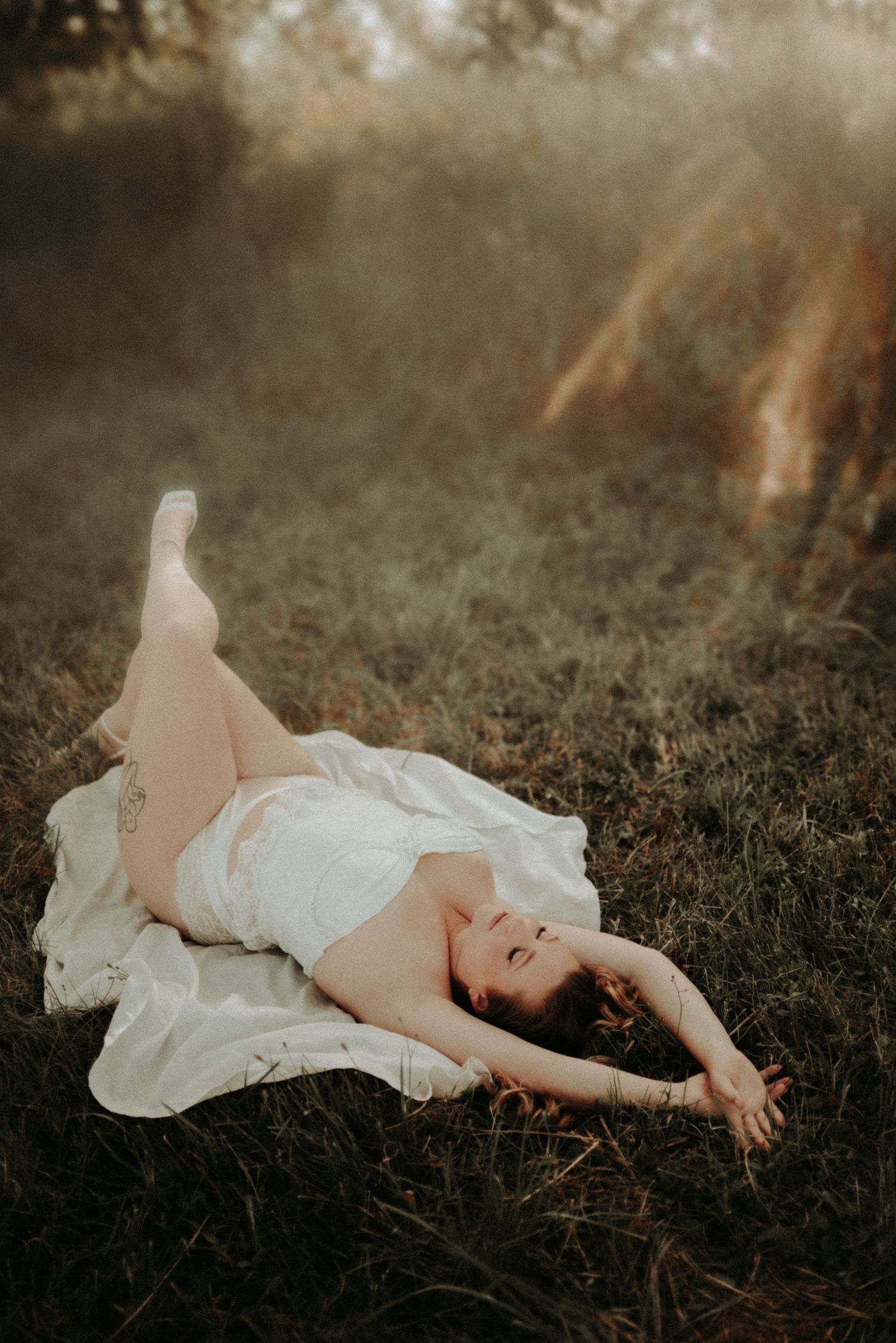 A person in white clothing rests peacefully on grass in a soft, artistic outdoor photoshoot.