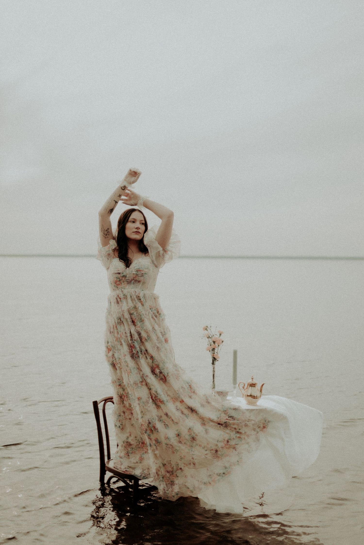 Figure in floral dress poses dramatically by water on misty day.