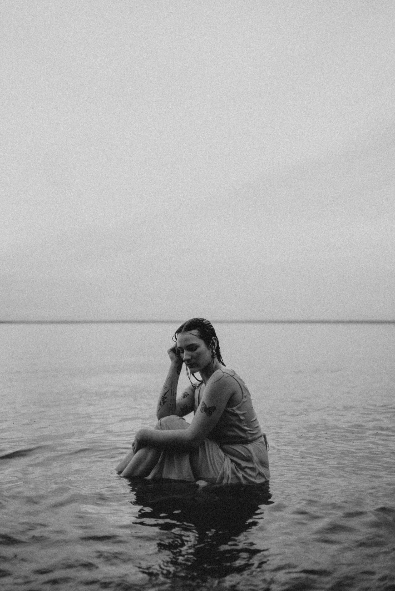 A black and white portrait of someone sitting thoughtfully in shallow water.
