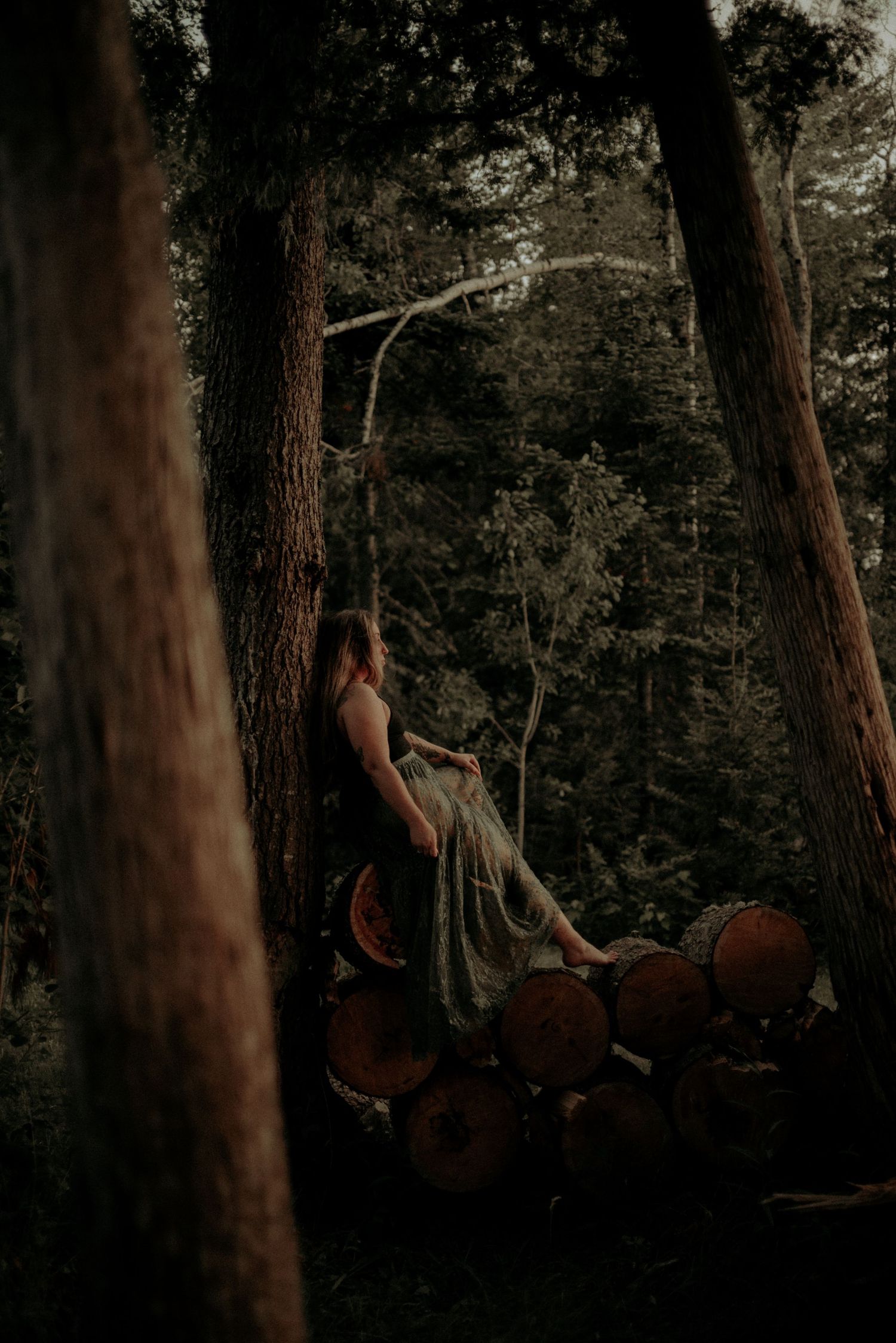 Figure in dark clothing perched on stacked logs in moody forest setting.