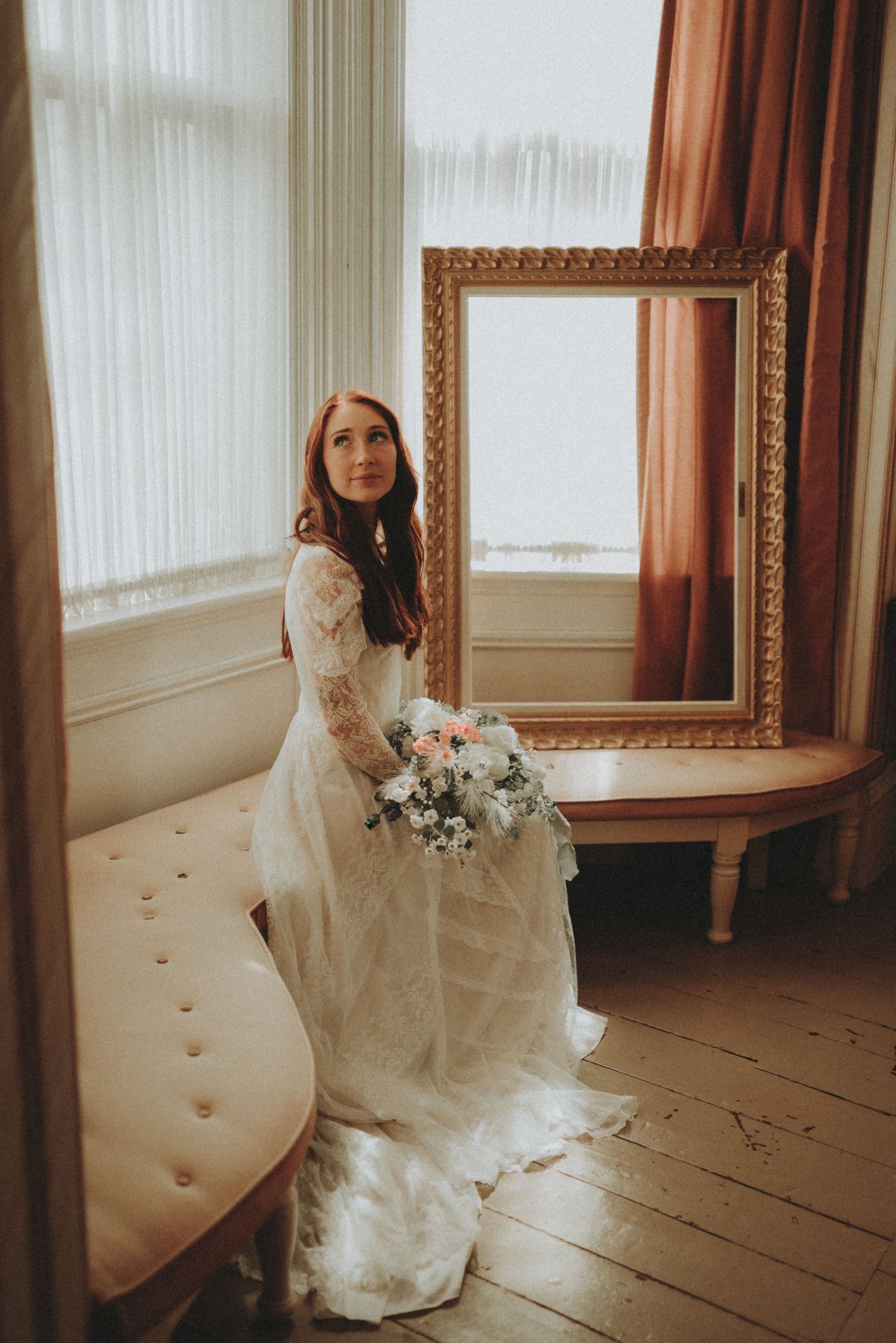 Romantic bridal portrait in a vintage room with lace wedding dress and delicate bouquet near an ornate gold mirror.