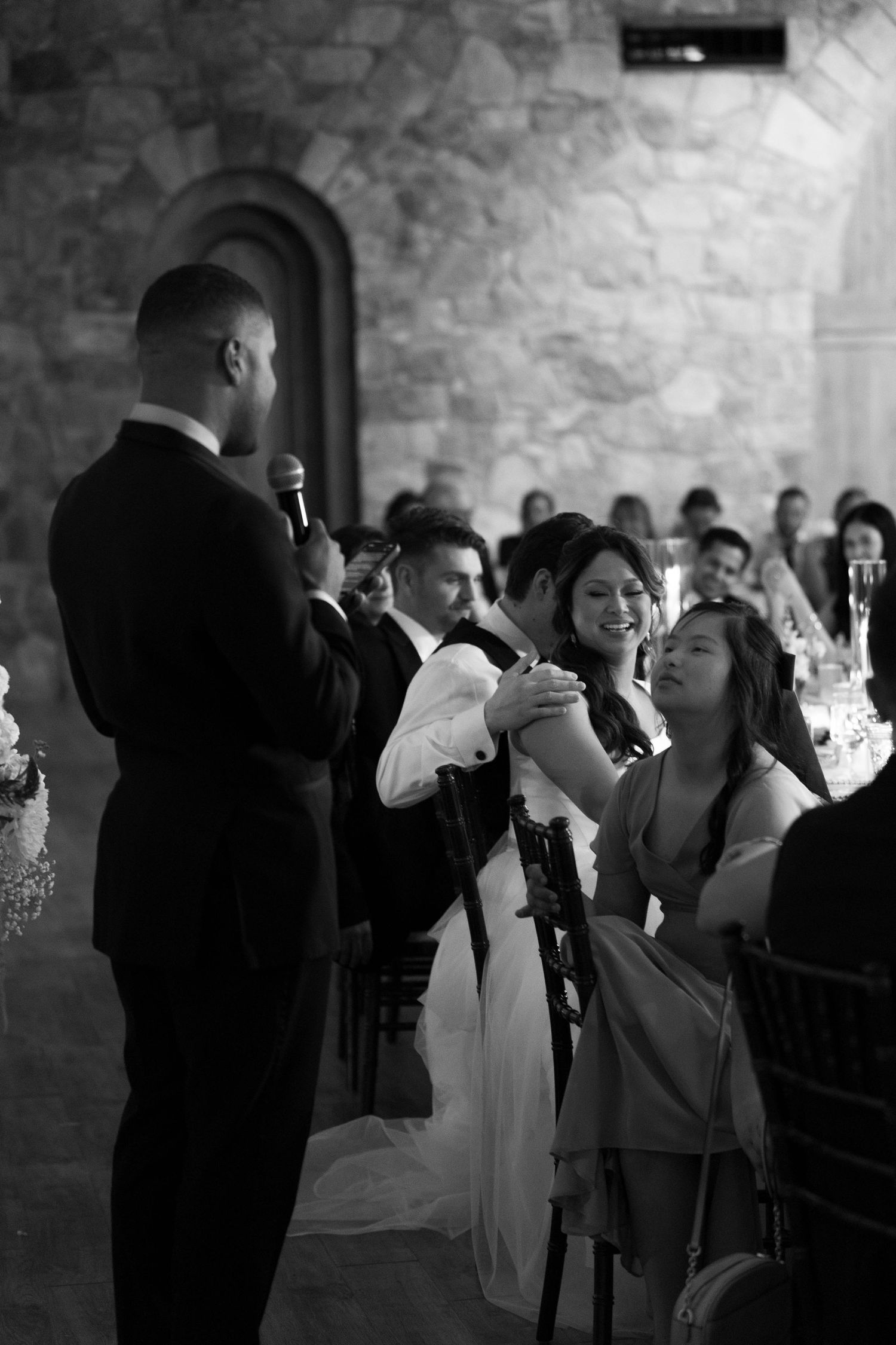 Black and white wedding reception photo of guests smiling as a man gives a heartfelt toast with a microphone.