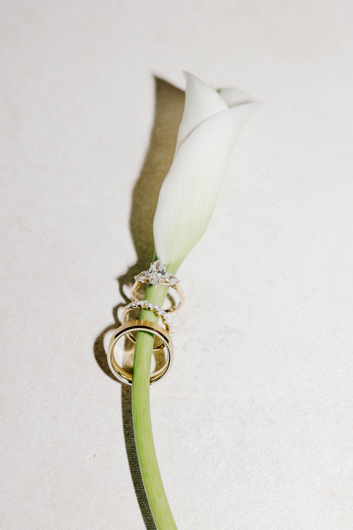 Three gold wedding rings stacked on a white calla lily stem against a light background.
