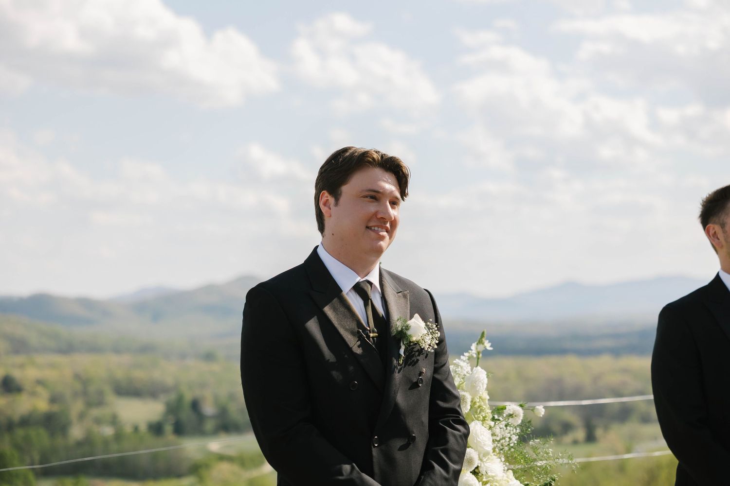 Smiling groom in black suit with white boutonniere at outdoor wedding ceremony with scenic mountain valley backdrop.