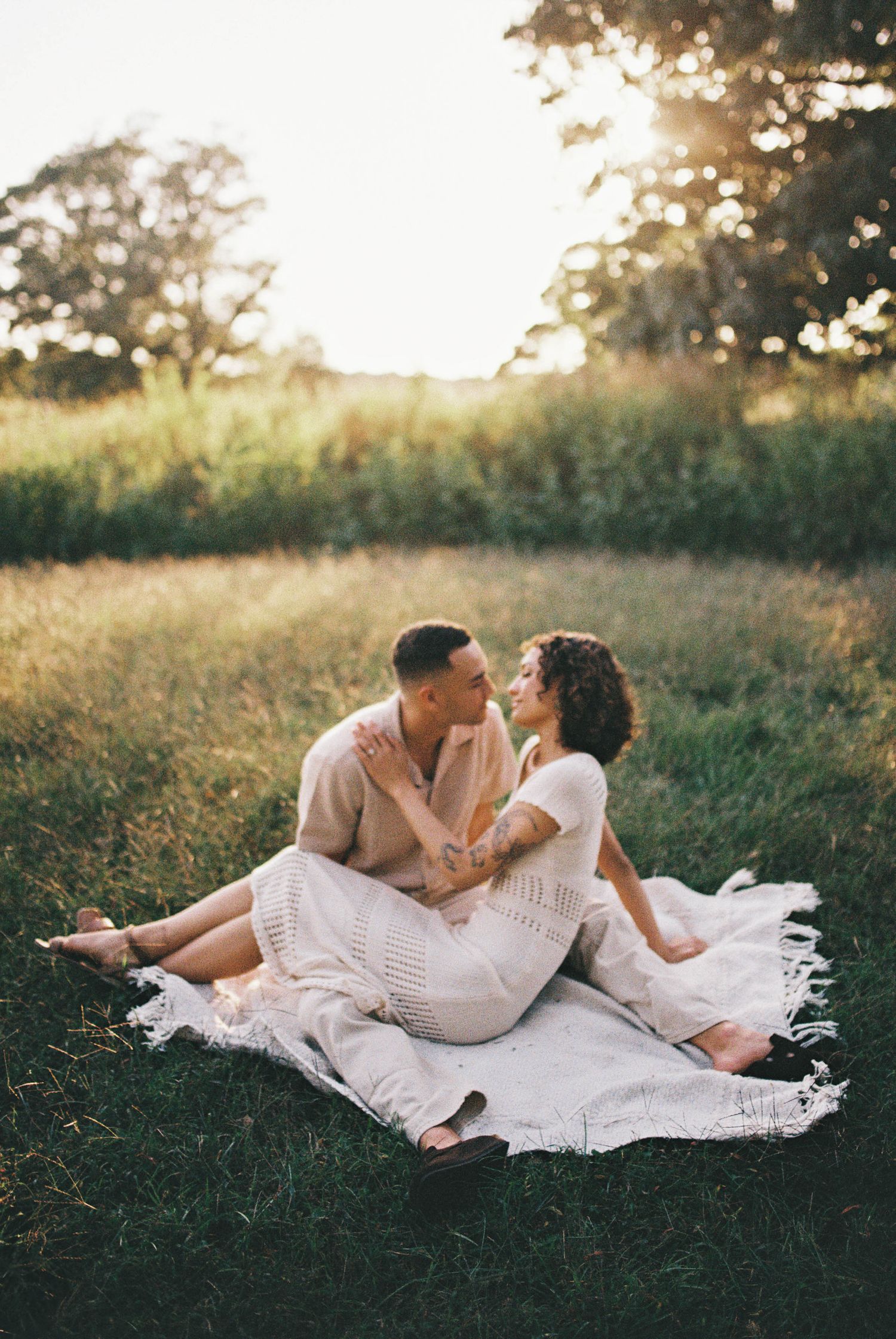 A couple in white outfits lying together on a blanket in a grassy field during golden hour sunset.