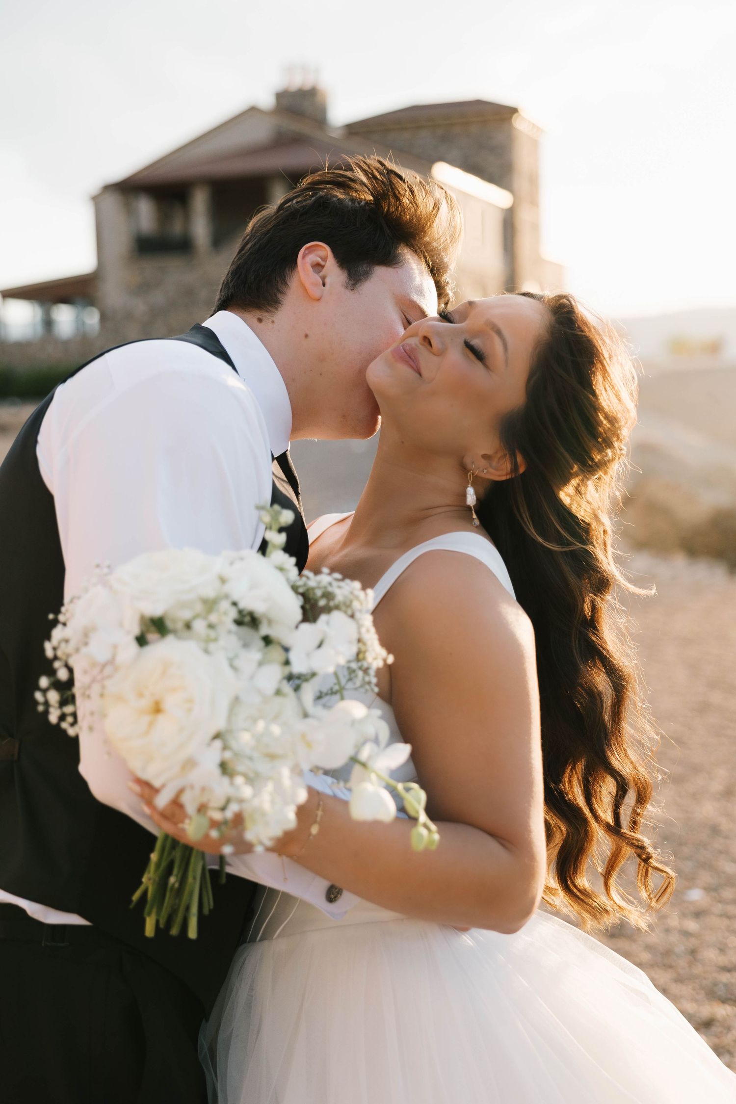 Groom kissing bride on cheek outdoors, bride holding white floral bouquet in elegant wedding attire.