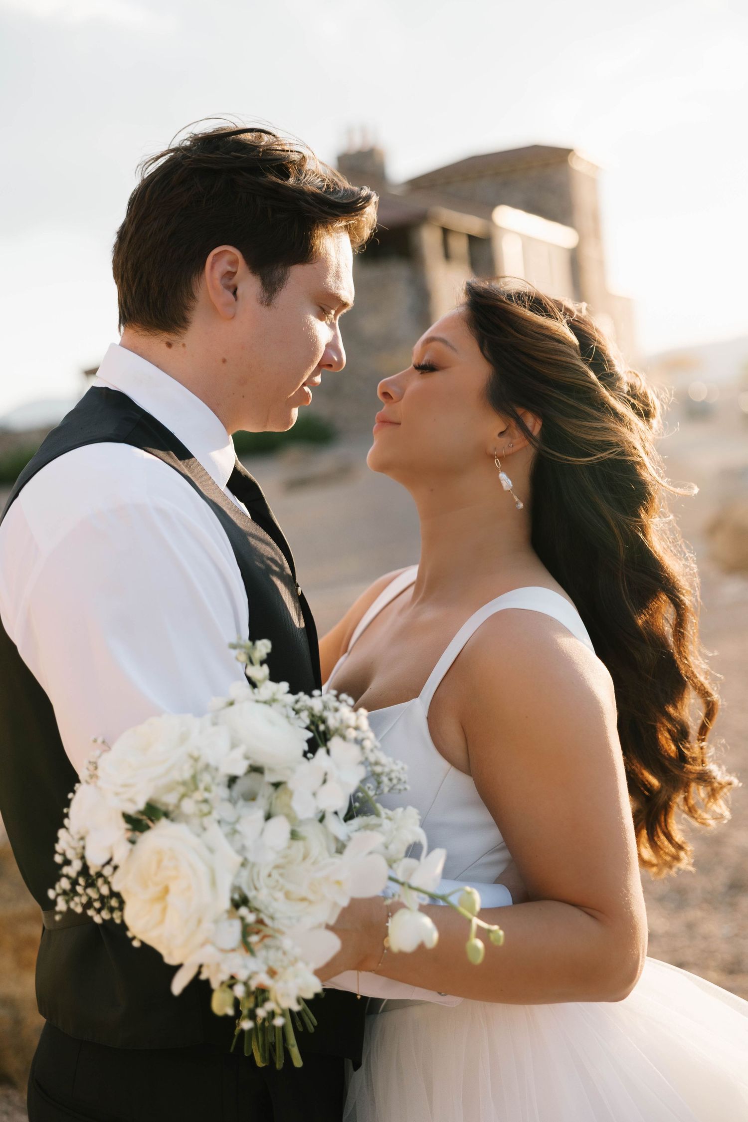 Bride and groom sharing a romantic moment outdoors, bride holding white floral bouquet in elegant wedding attire.