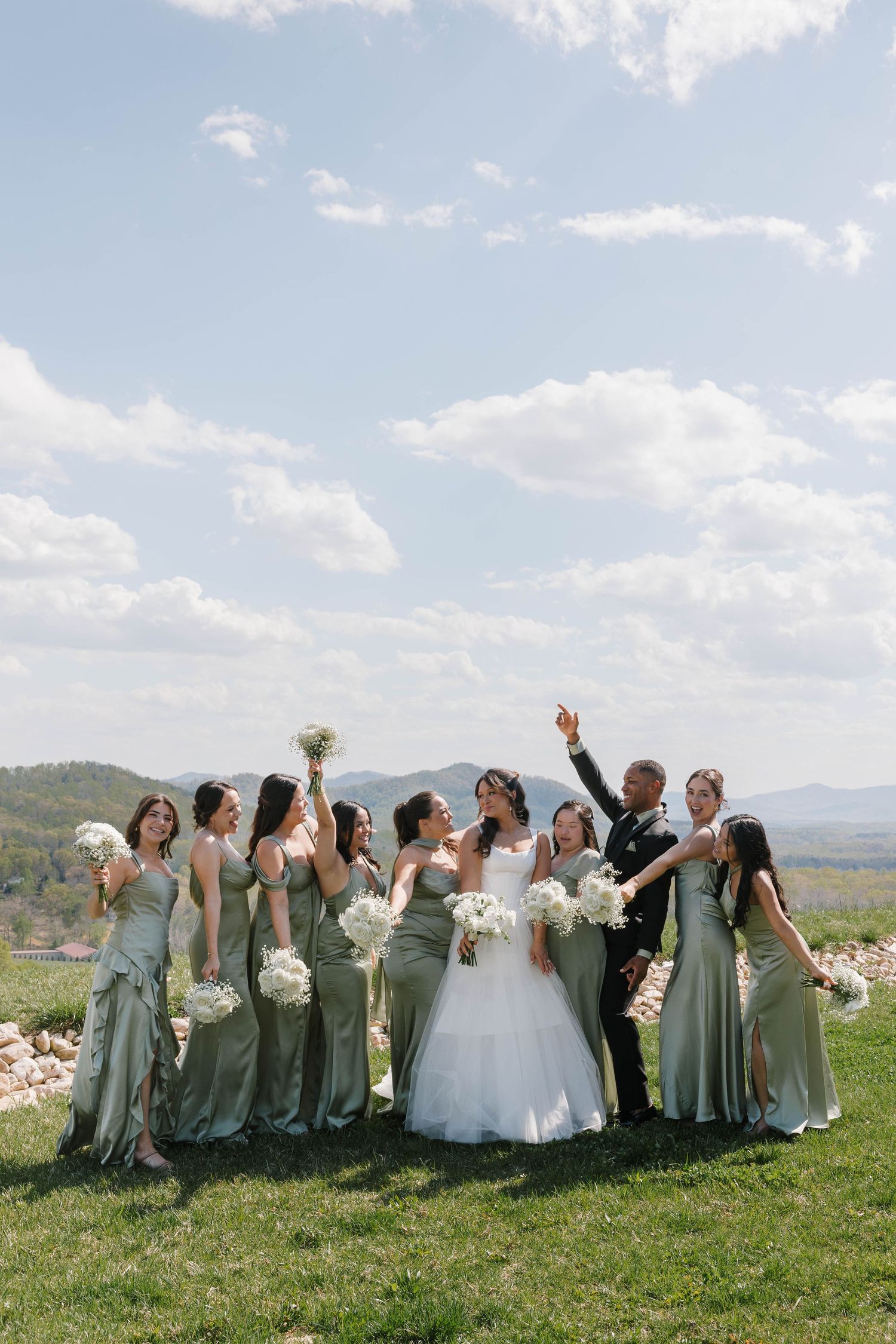 Bride and groom celebrate with bridesmaids in sage green dresses, holding white bouquets outdoors with mountain views.