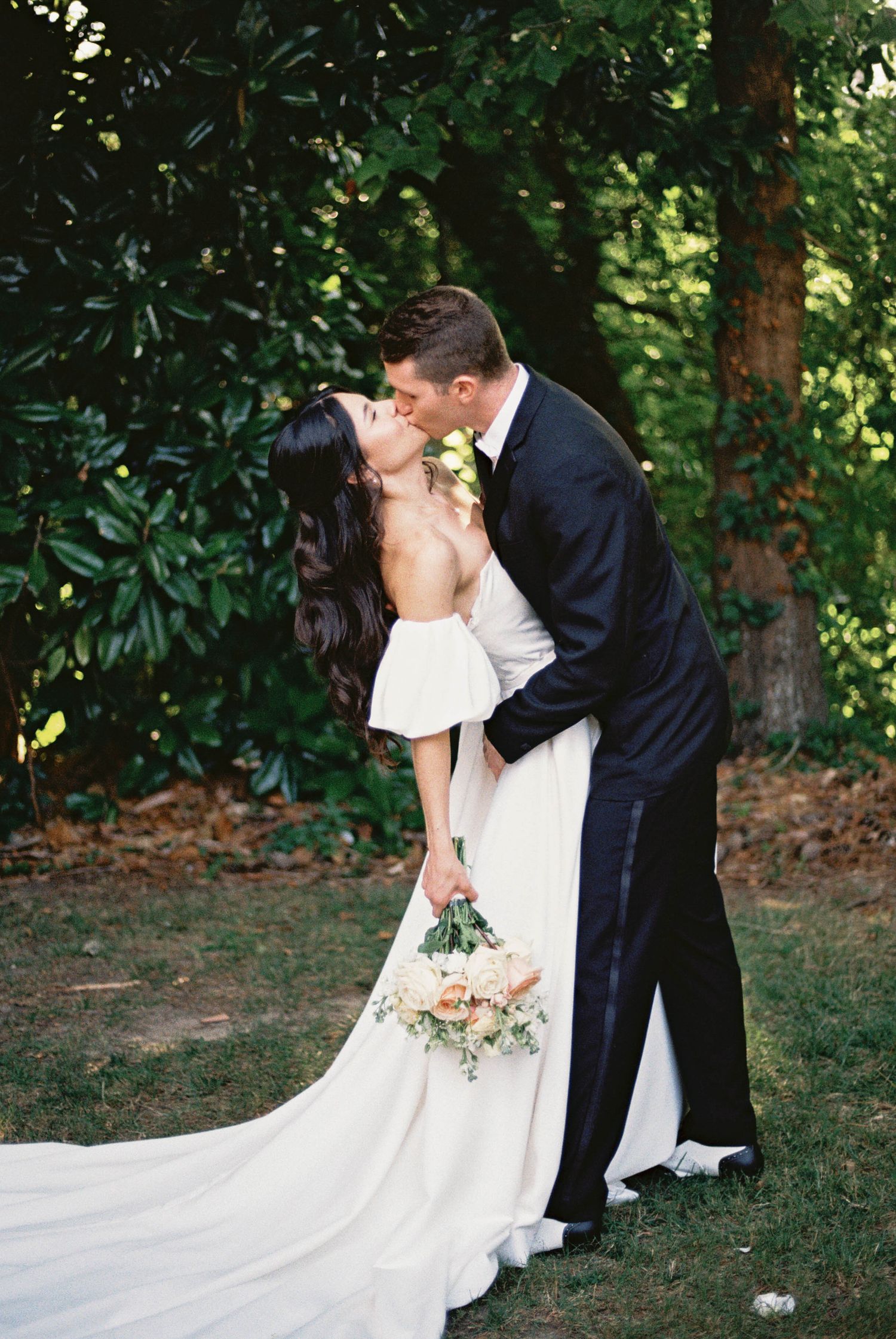 A wedding couple shares a kiss in a garden setting with lush greenery and a flowing white bridal gown at woman's club of portsmouth