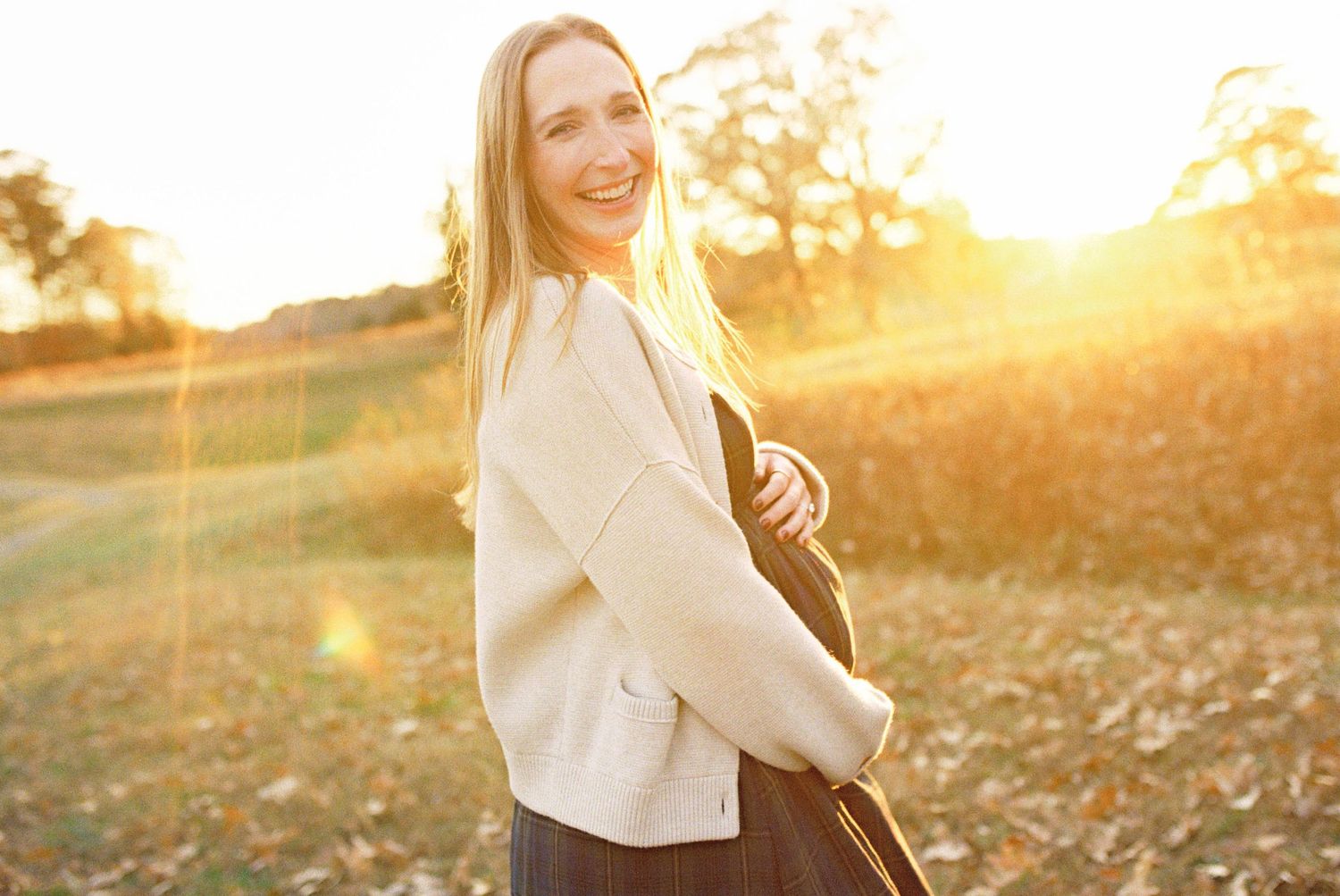 Person in white sweater poses in autumn field during golden hour sunset with sunflare and fall foliage.
