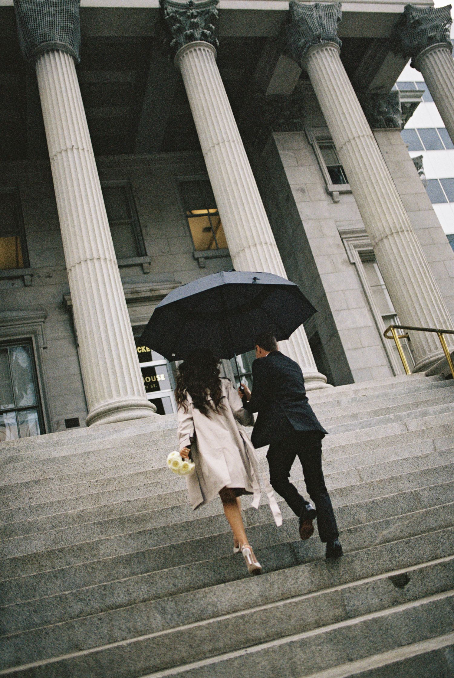 Couple running up stairs under umbrella near classical columns in the rain.