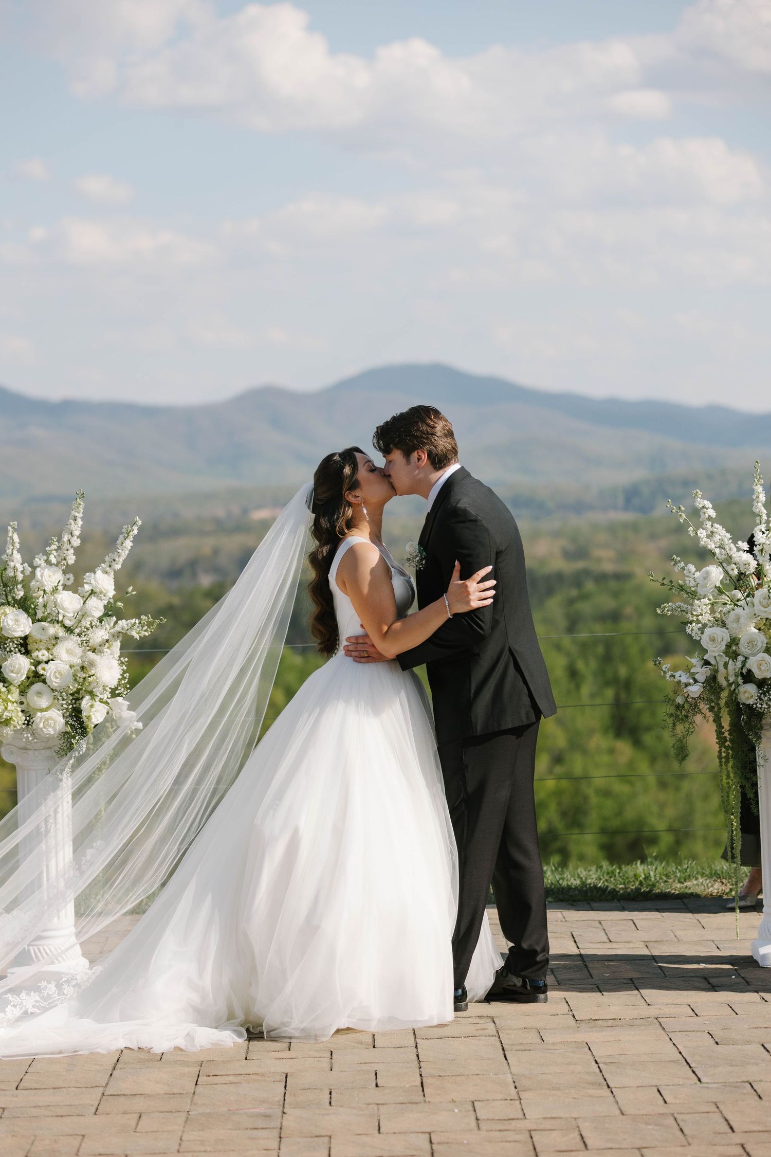 Bride and groom share first kiss at outdoor wedding ceremony with stunning mountain landscape backdrop and white floral arrangements.