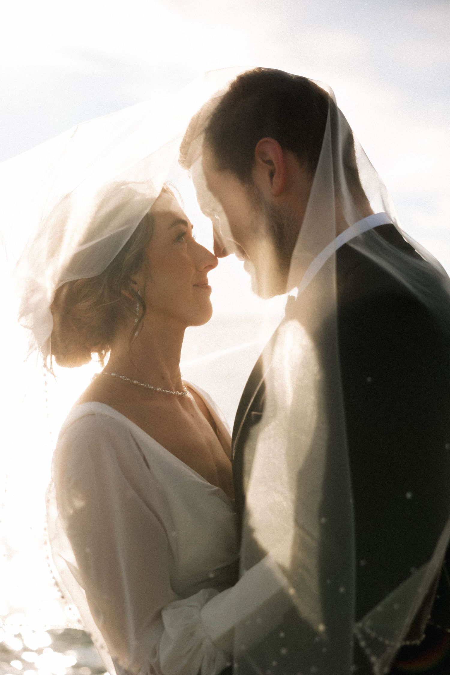 Backlit wedding couple in ethereal white veil creating romantic silhouette.