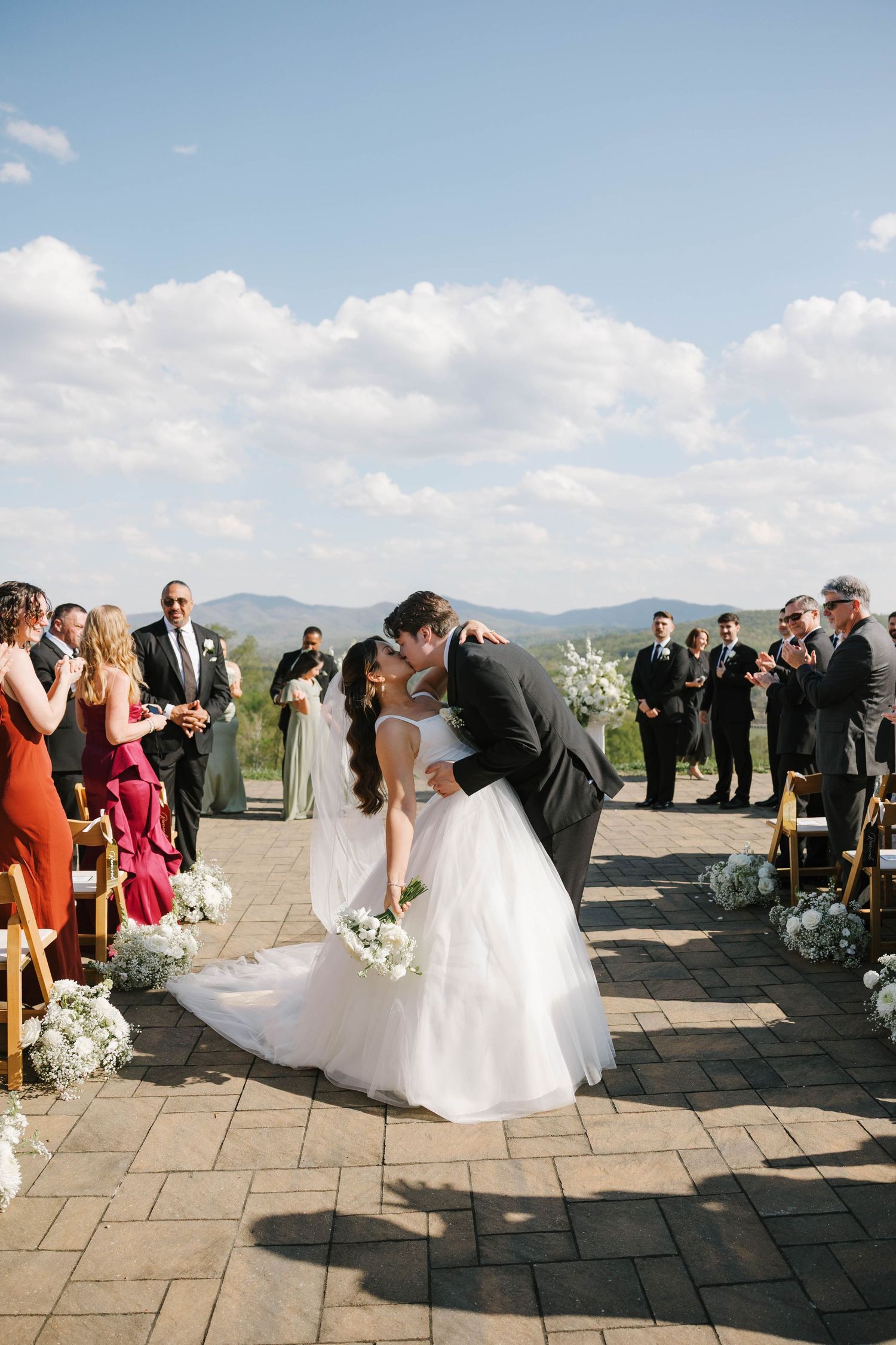 Bride and groom share first kiss at outdoor wedding ceremony with mountain views and guests applauding.