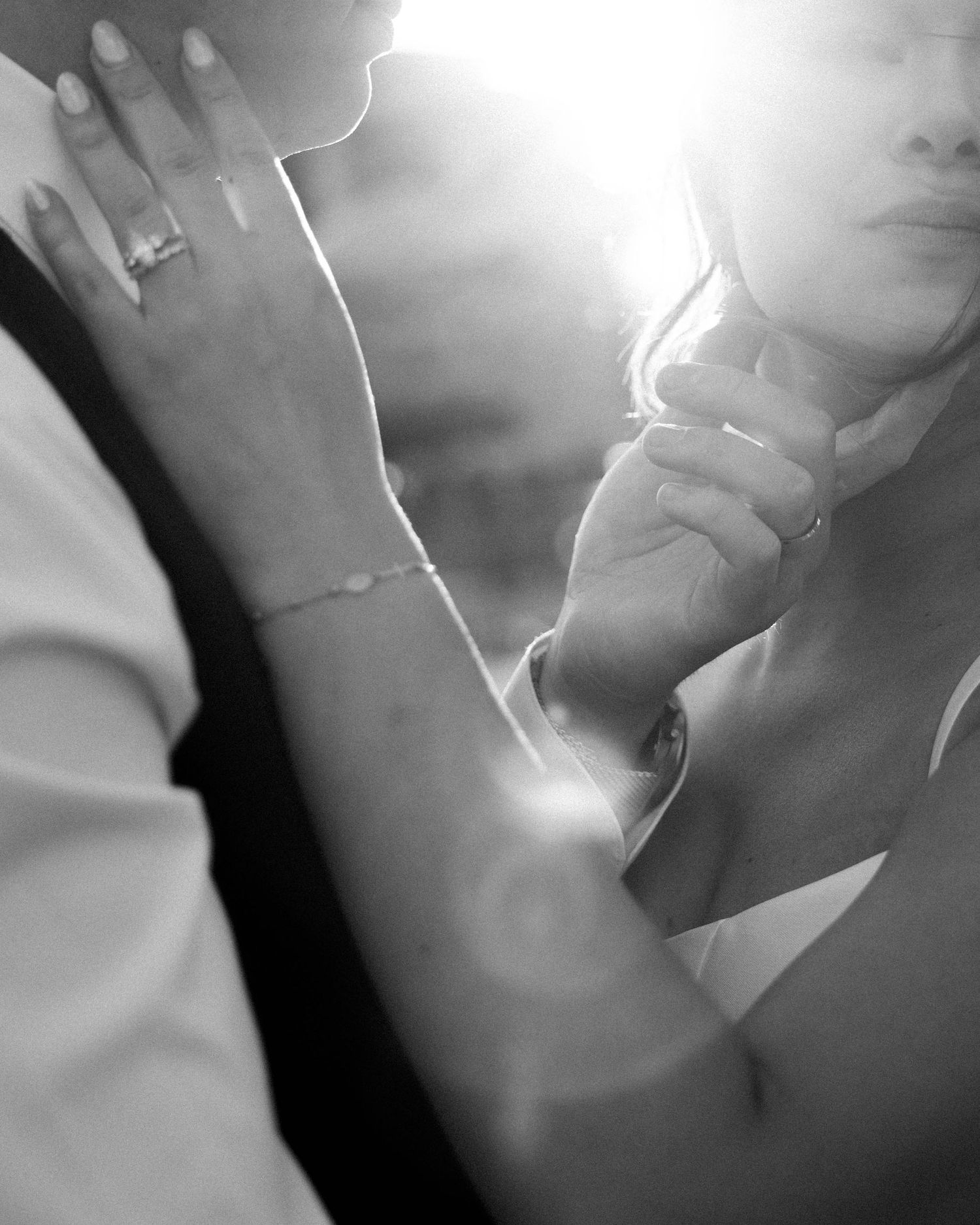 Black and white wedding photo of bride and groom holding hands, engagement ring and bracelet visible, backlit with soft glow.