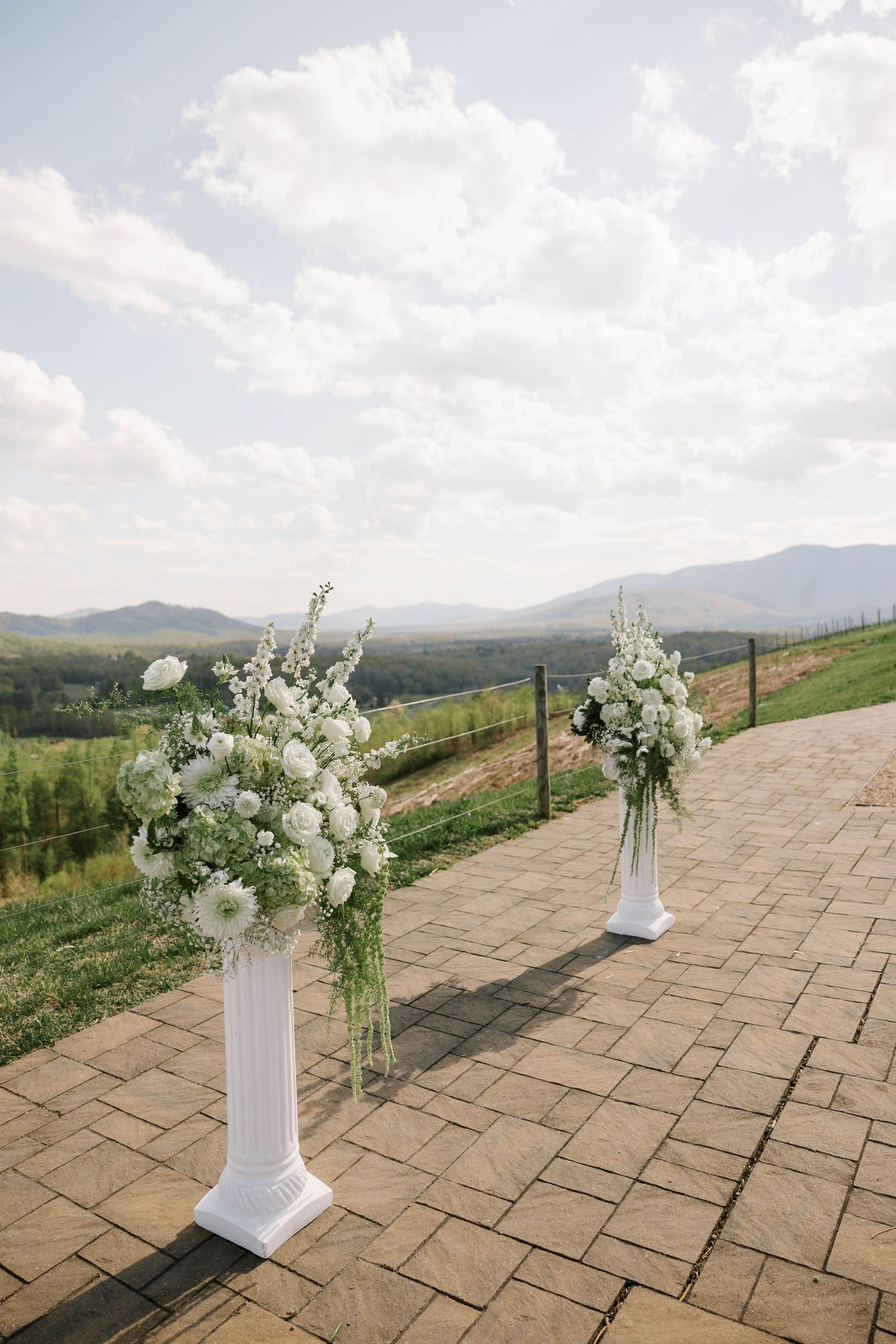 Two white floral pedestals with elegant white flower arrangements on a brick patio overlooking scenic mountain views.