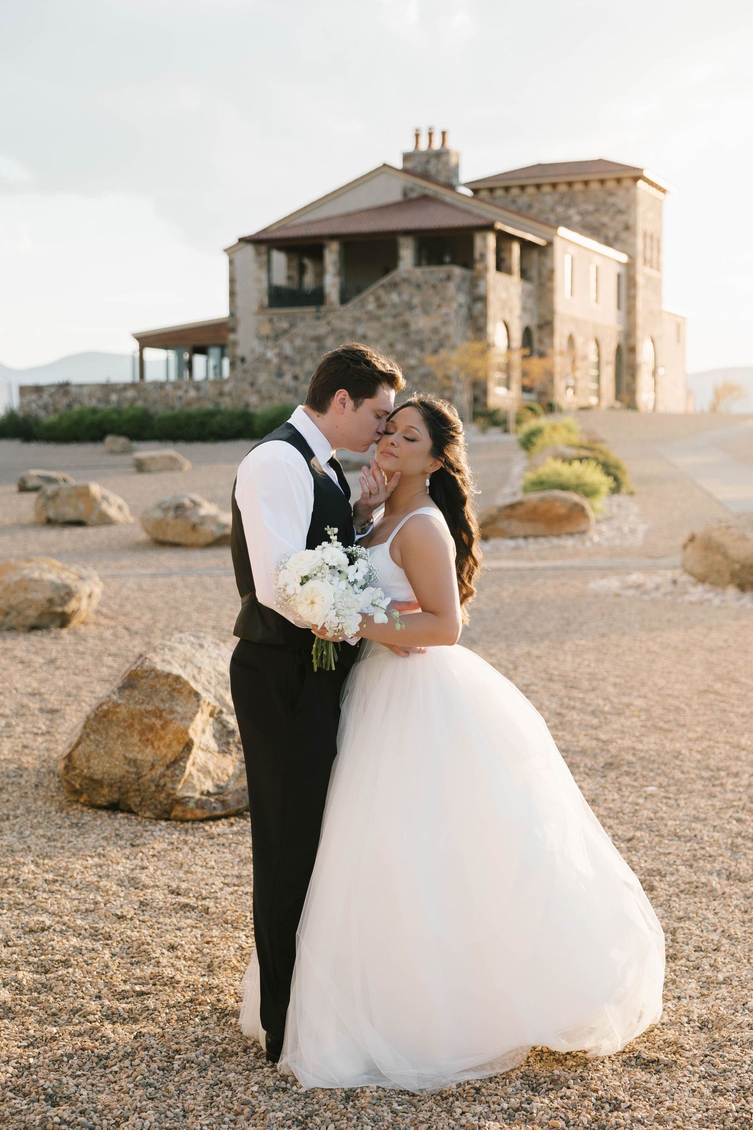 Bride in white ballgown and groom in black tuxedo embrace outdoors near stone mansion at golden hour.