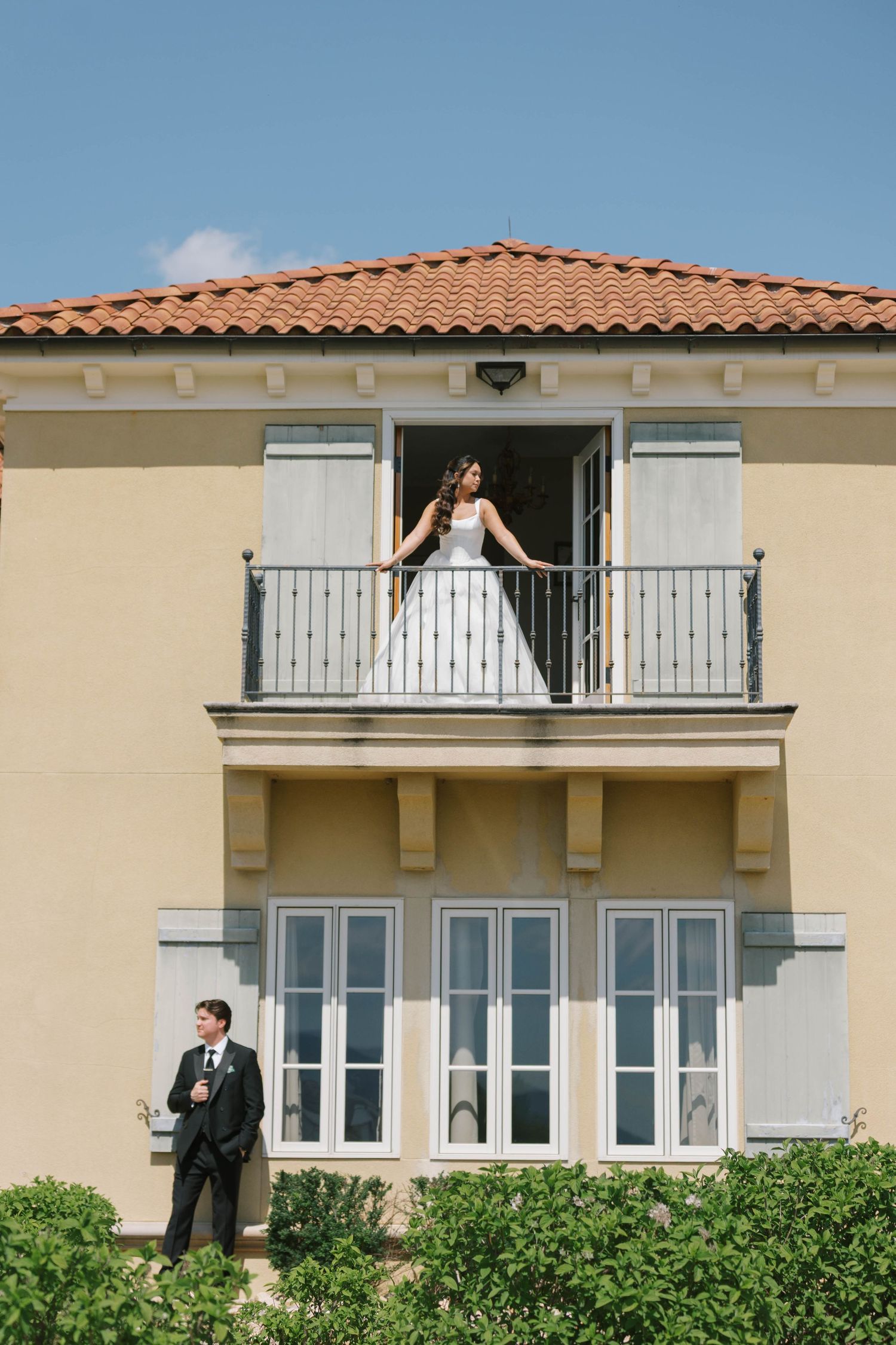 Bride in white gown on balcony with groom below at elegant yellow villa wedding venue.