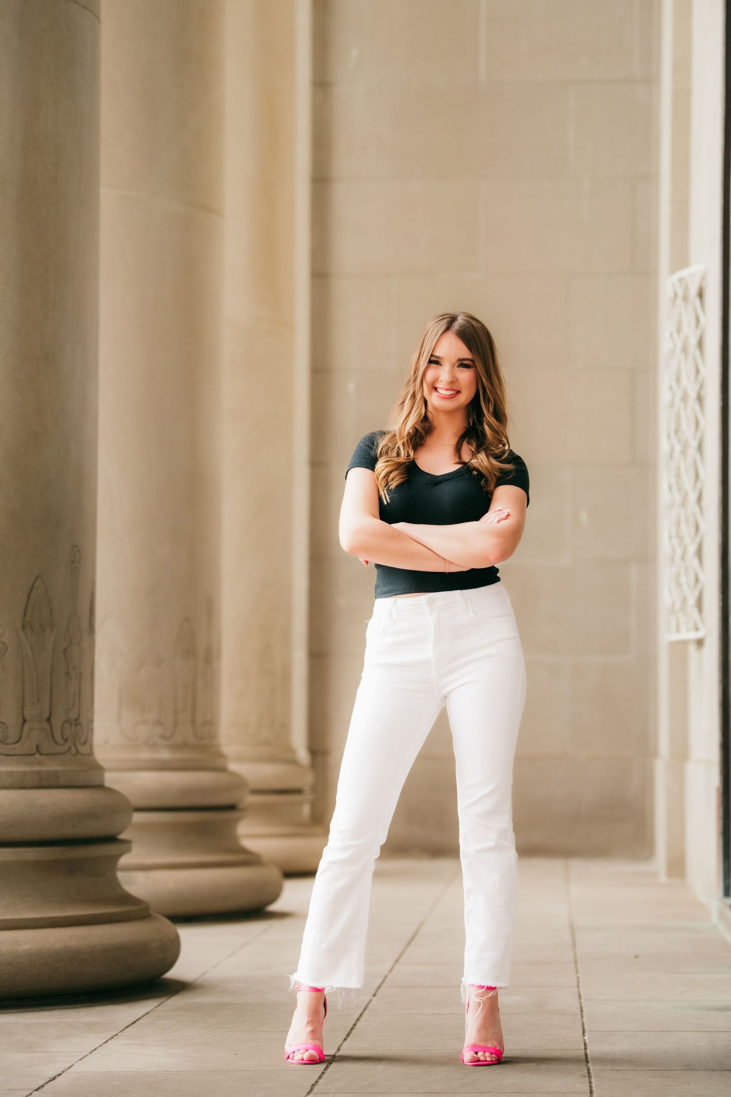 Woman in black top and white pants poses elegantly between classical stone columns.