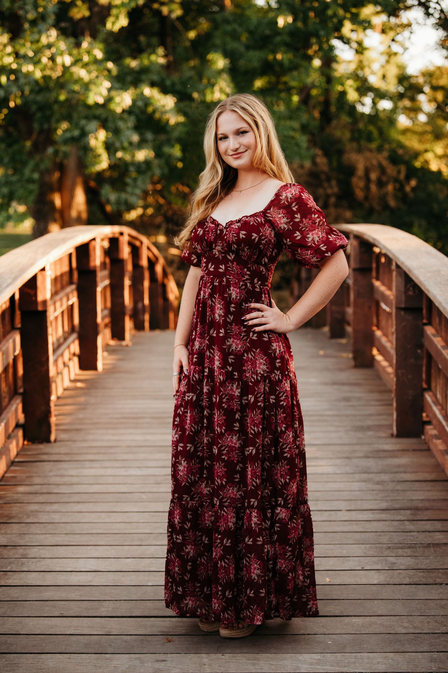 A woman in a burgundy floral dress poses on a wooden bridge during golden hour sunset.