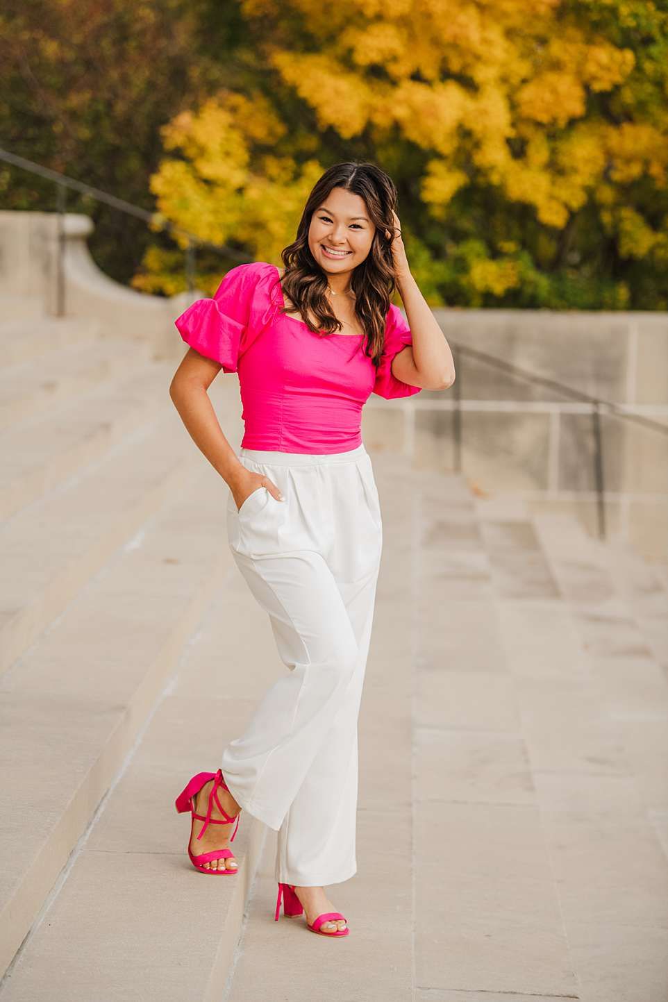 Fashion shoot with bright pink top and white pants against autumn foliage backdrop.