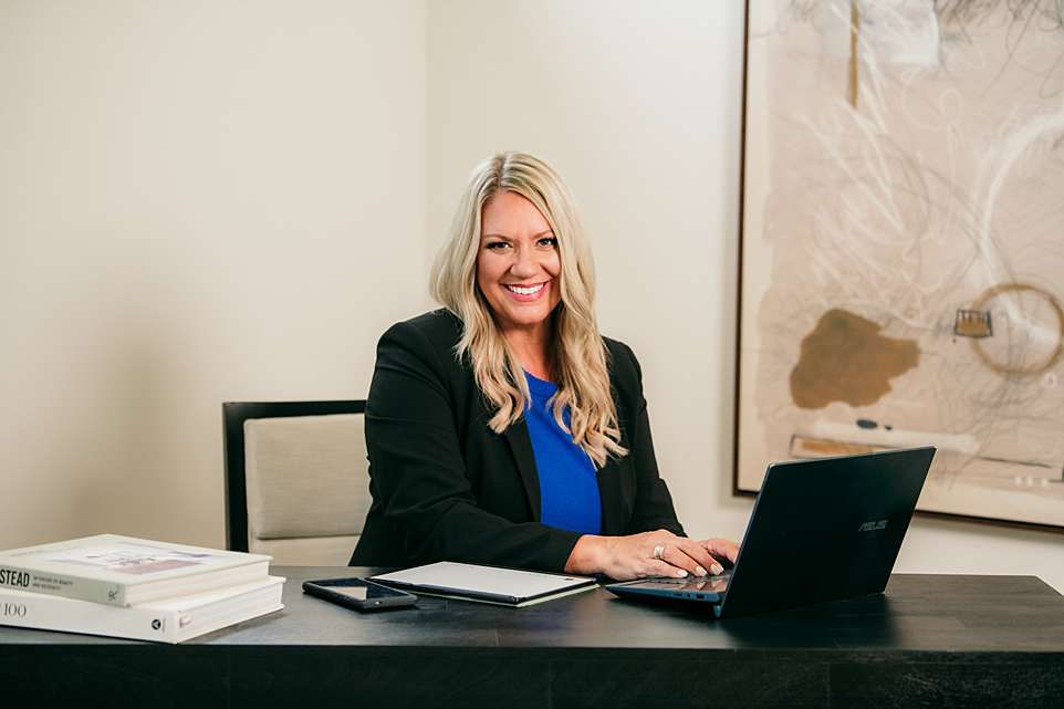 Woman in business attire smiles while working at desk with laptop in bright office setting.