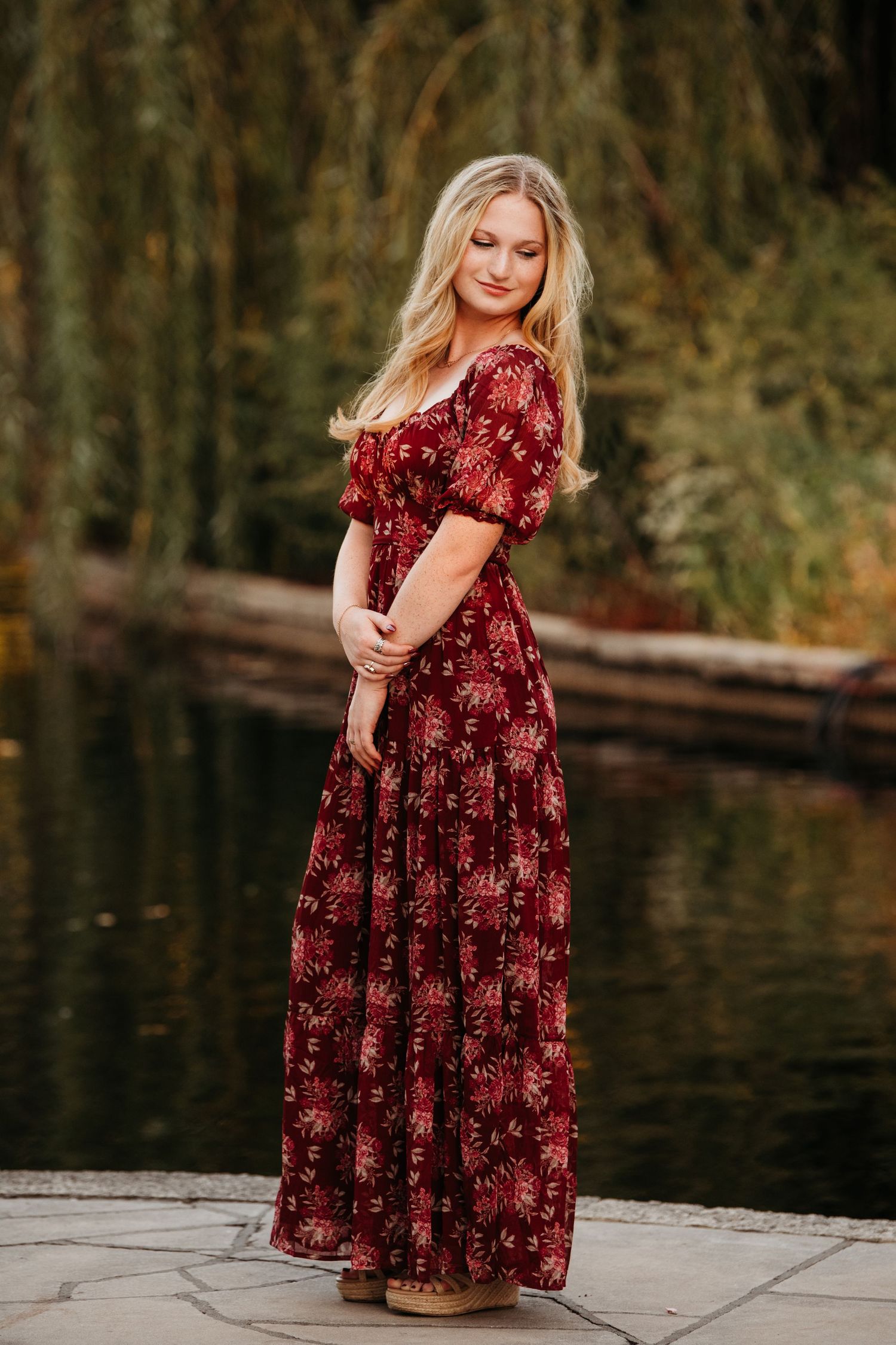 A person in a burgundy floral dress stands on a wooden dock by a serene lake during autumn.