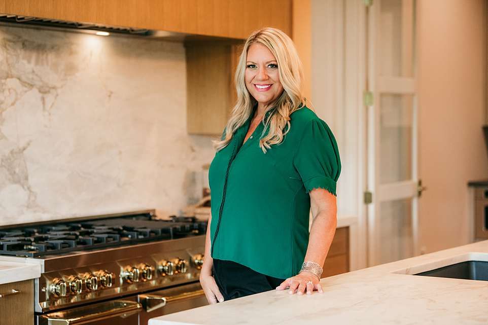 Woman in emerald green top stands in modern kitchen with stainless steel appliances and white cabinets.