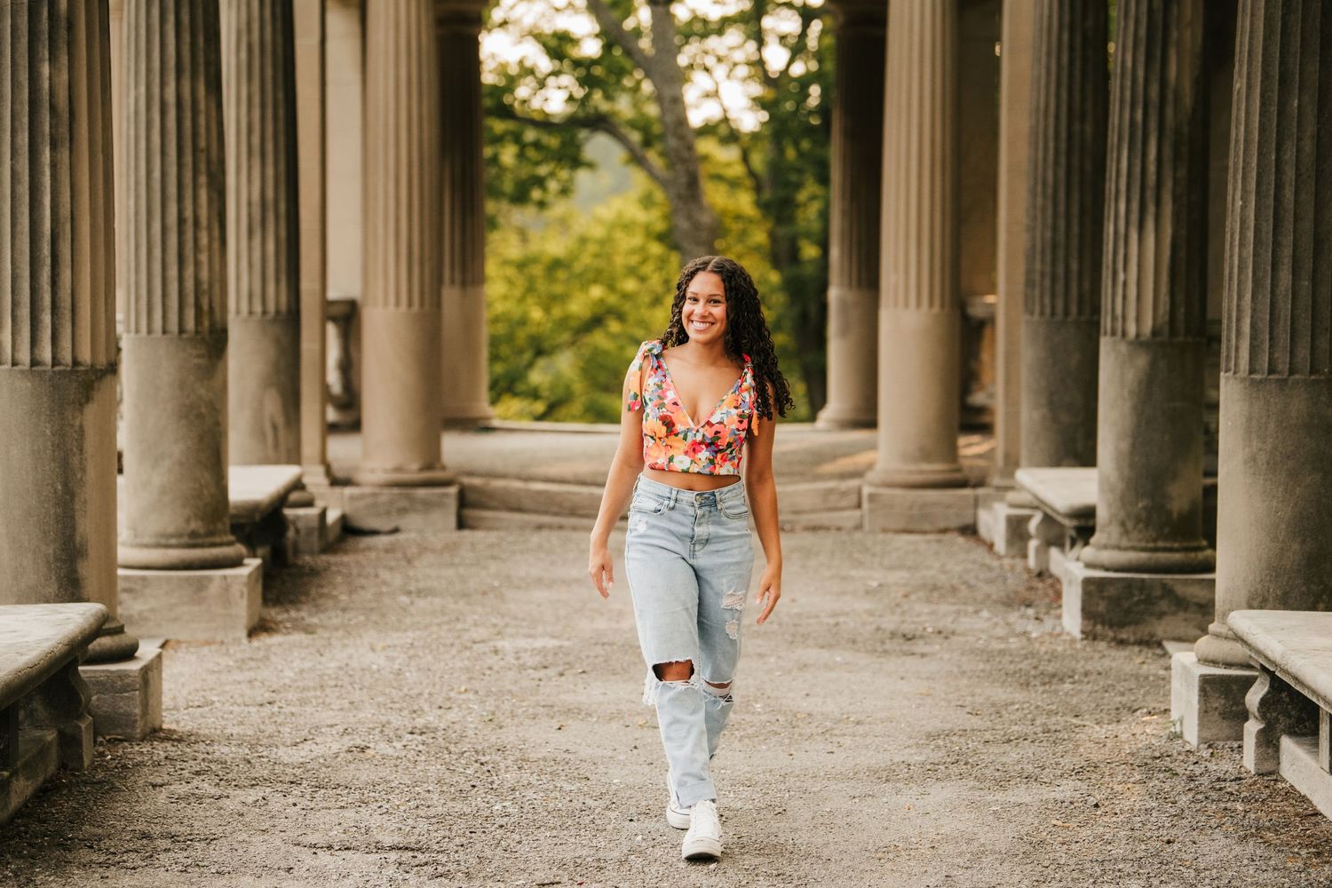Someone walks through a covered outdoor colonnade pathway with stone columns and gravel ground on a sunny day.