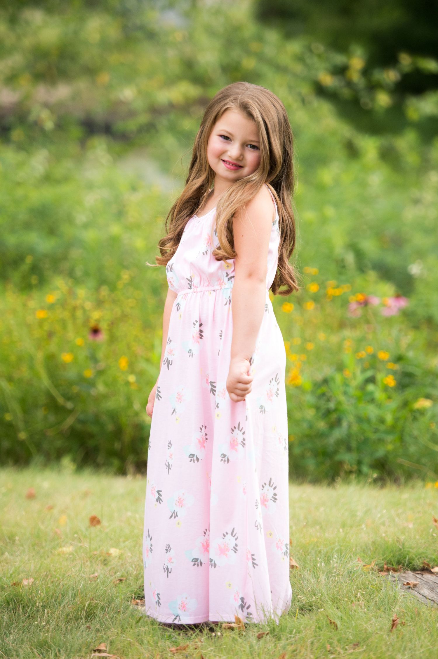 A young child wearing a long pink floral dress stands in a garden with wildflowers and greenery in the background.
