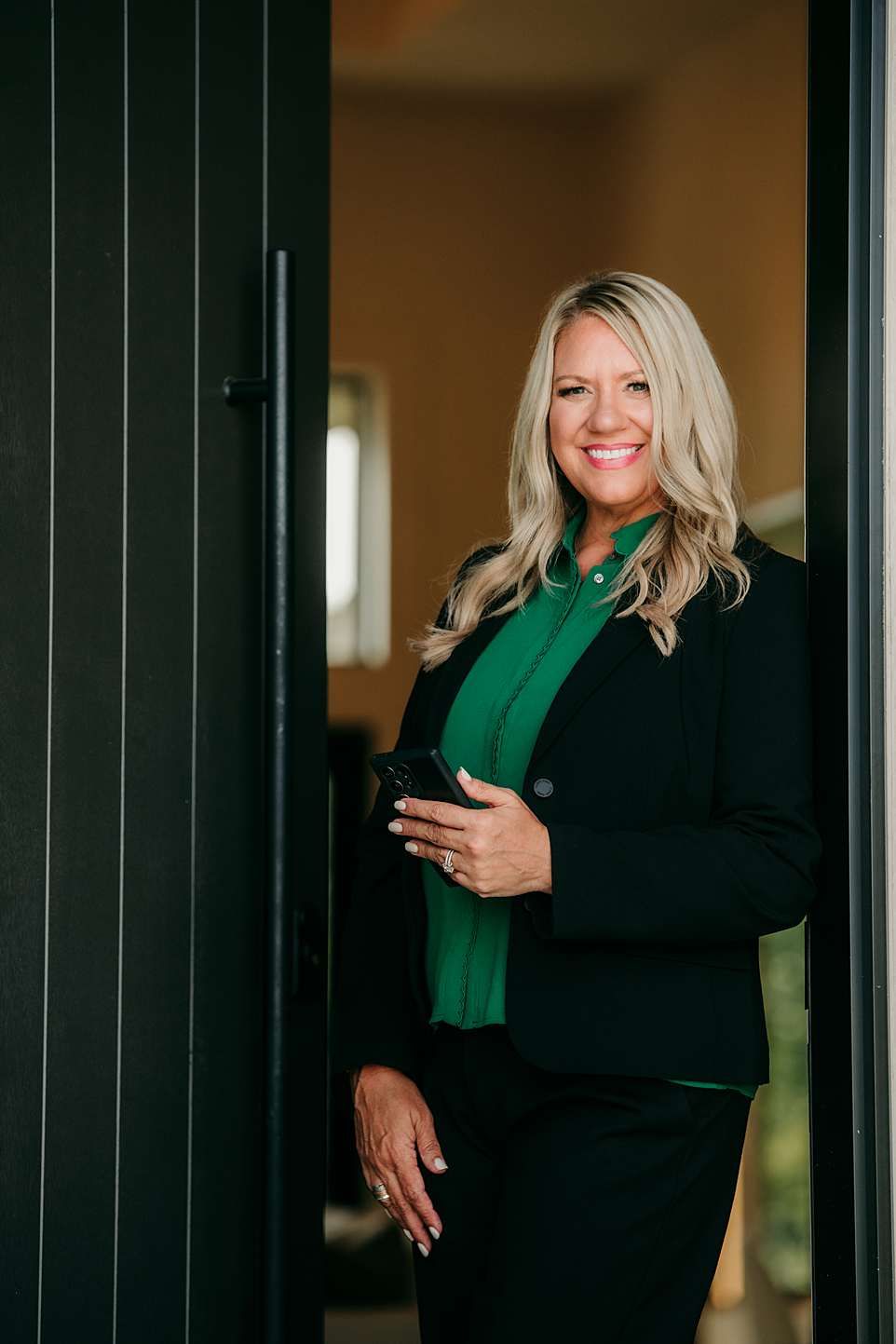 Woman in black blazer and green top poses against dark wall for professional portraits.