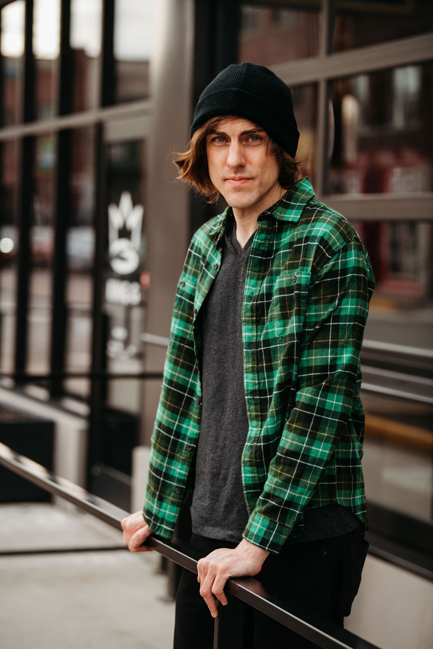 Urban portrait featuring green plaid flannel jacket and beanie against modern building backdrop.