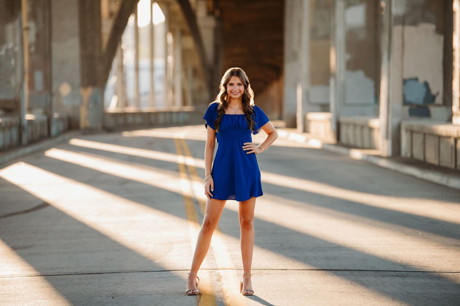 Woman in blue dress poses elegantly in front of sunlit columns and architecture.