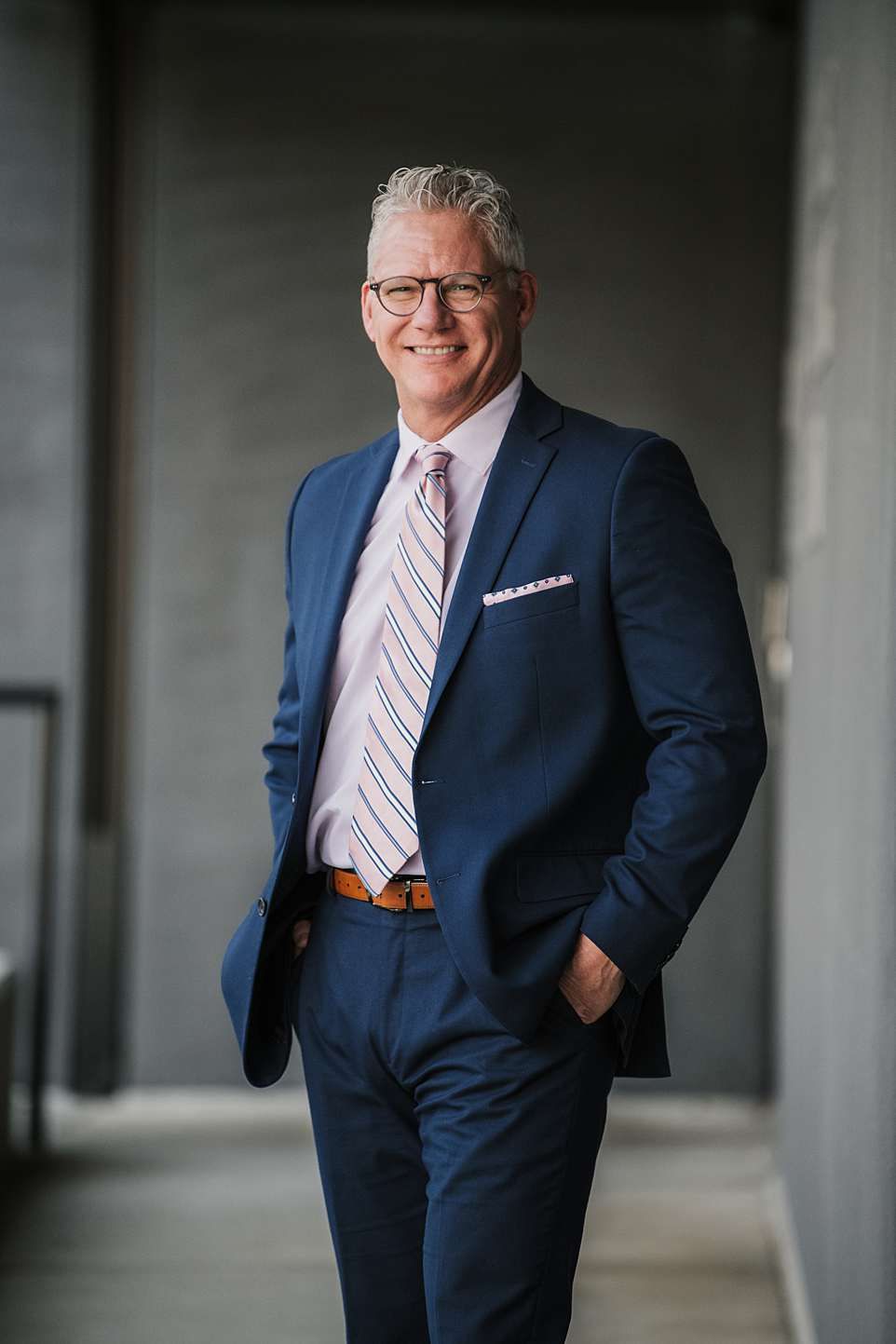 Professional business portrait of someone in navy suit and striped tie against grey background.