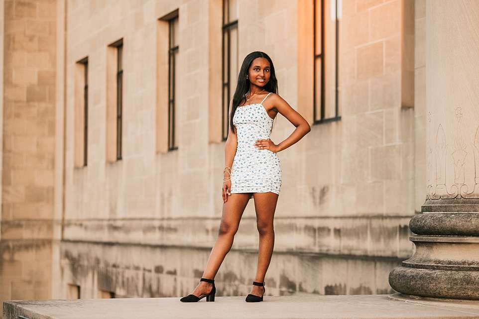 Full length portrait of person in white mini dress posing against stone building exterior.
