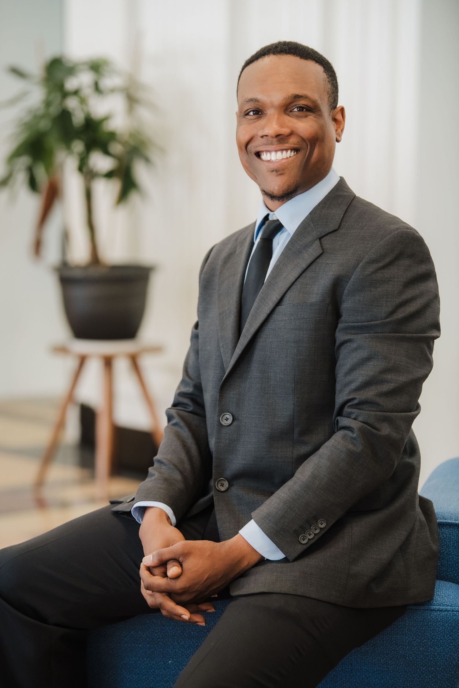 Professional headshots of someone in a gray suit sitting on a blue couch in a bright modern office setting.