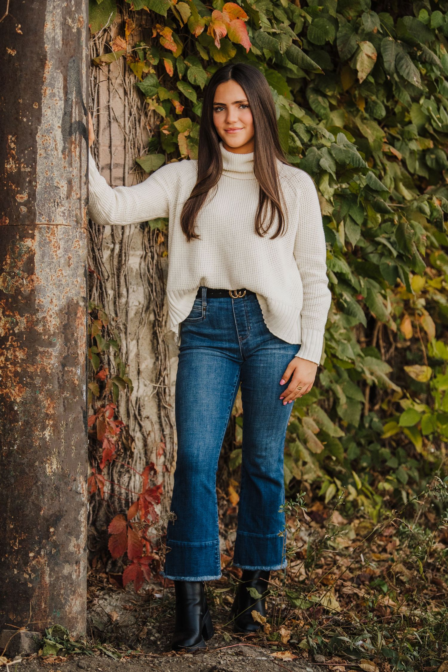 Person wearing white turtleneck sweater and blue jeans poses against rustic wall with autumn foliage background.