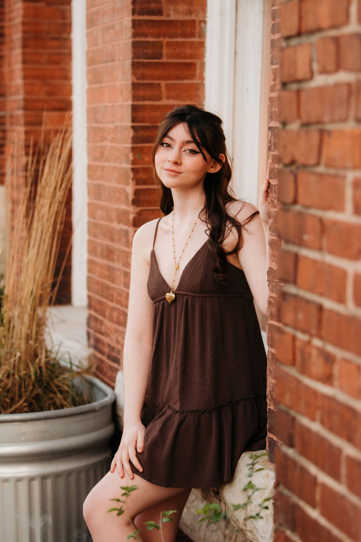 Portrait session against red brick wall with brown dress and decorative planter in natural lighting.