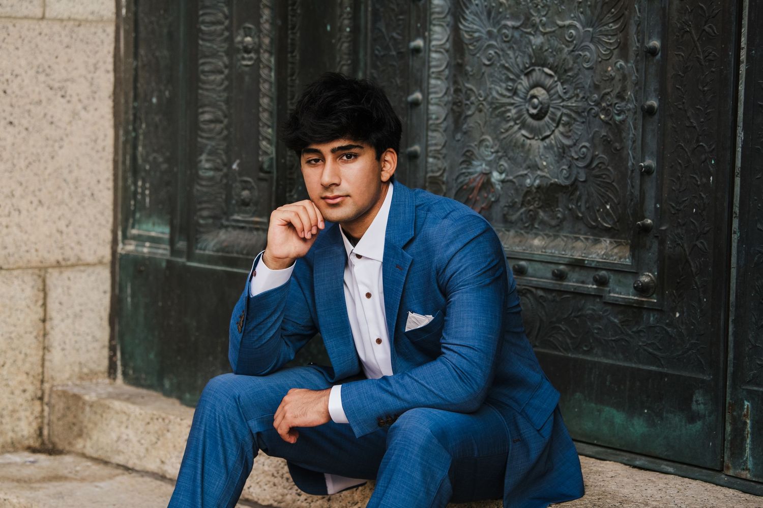 Person in navy blue suit posing confidently against stone wall backdrop.