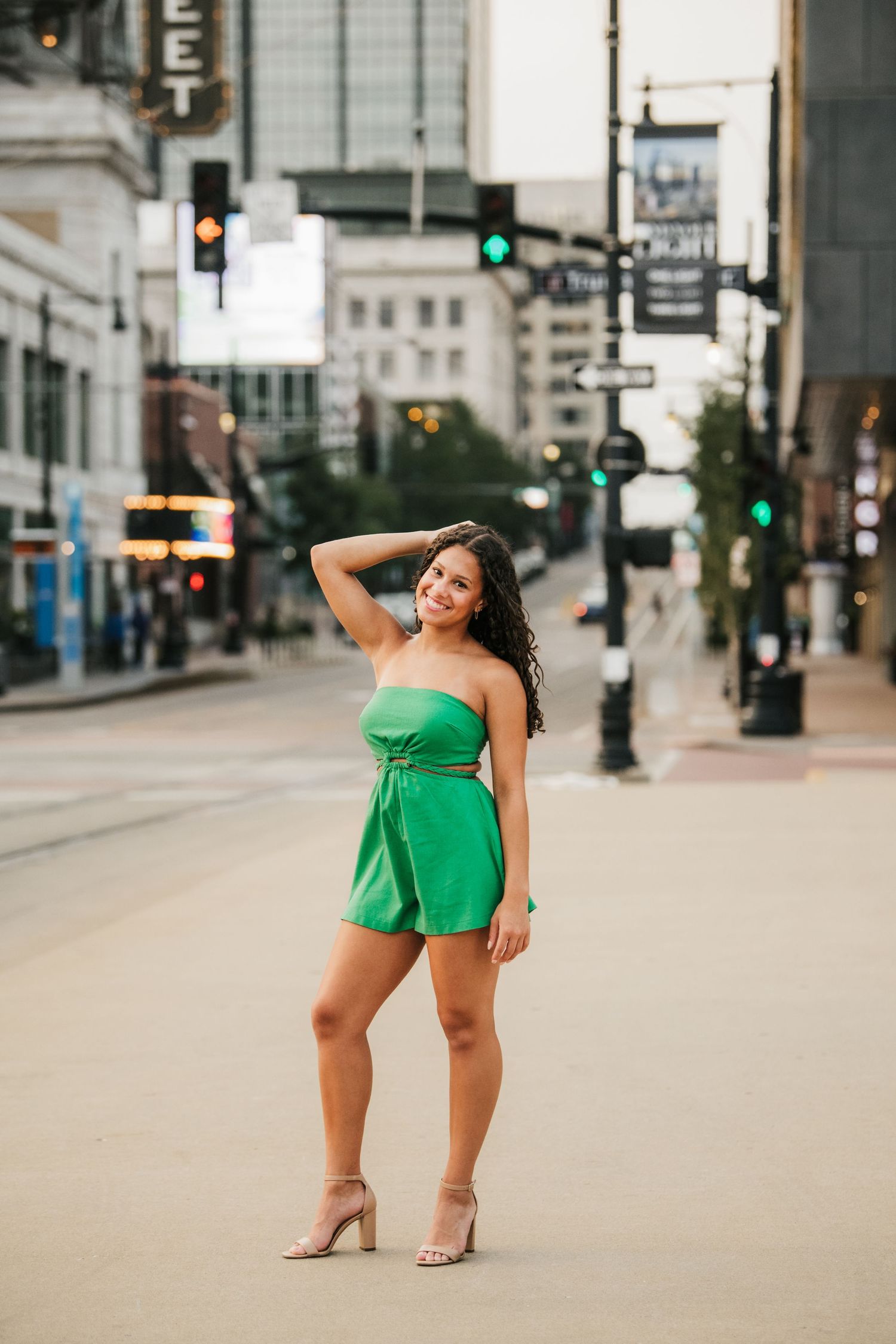 Woman in green outfit poses playfully on empty city street with buildings and traffic lights.