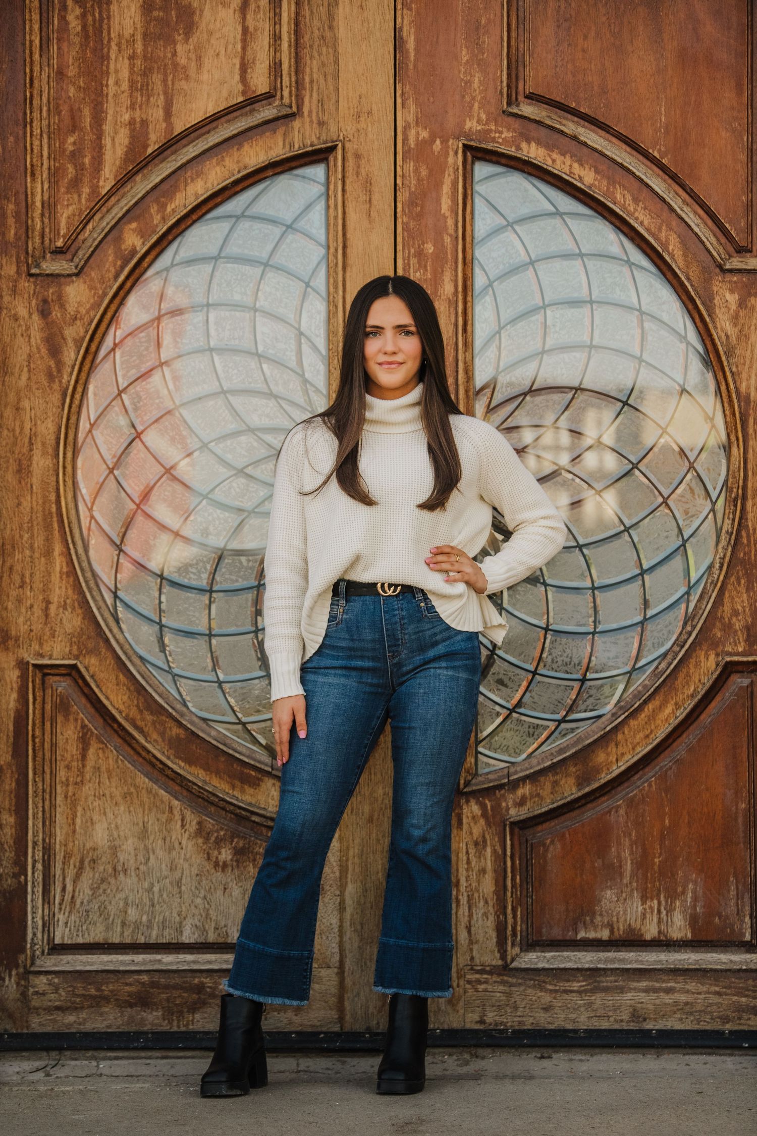 A person in a white turtleneck sweater and flared jeans poses in front of a vintage wooden door with circular glass panels.