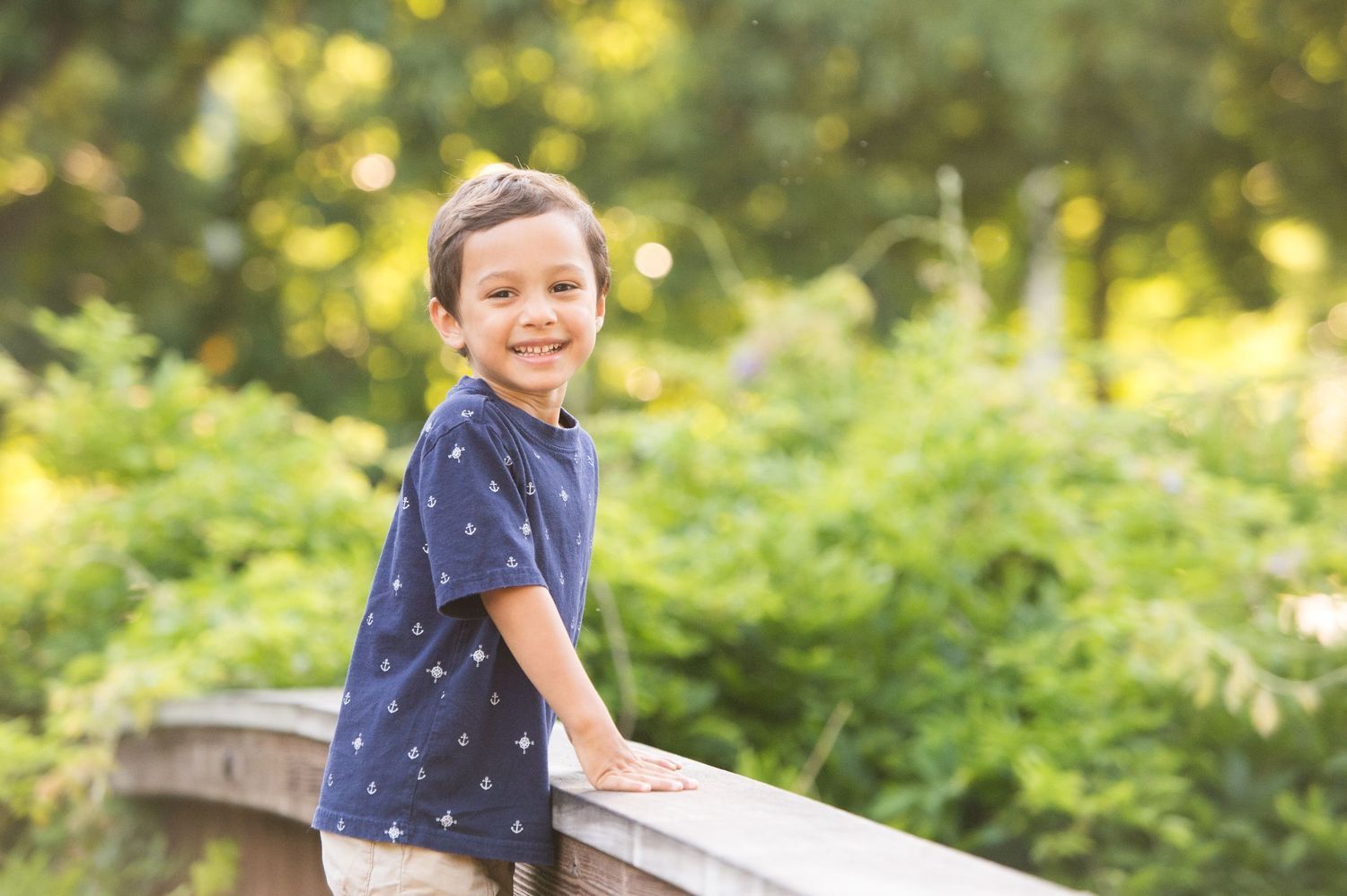 A child in a blue polka dot shirt leans against a wooden railing in a sunny garden setting.