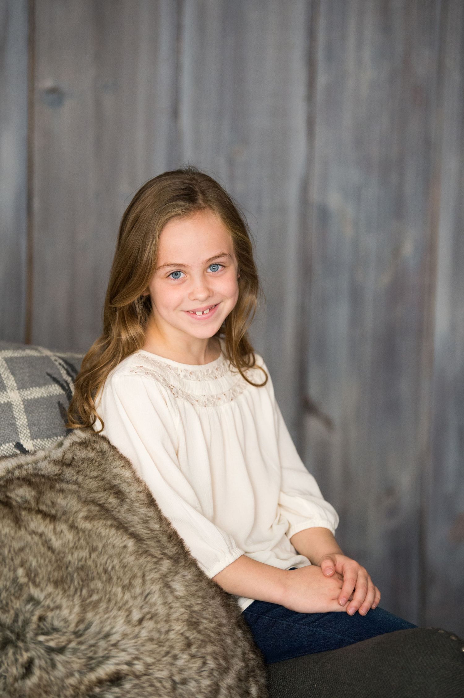 A young person in a white blouse sits on a gray couch while smiling at the camera in a studio setting.