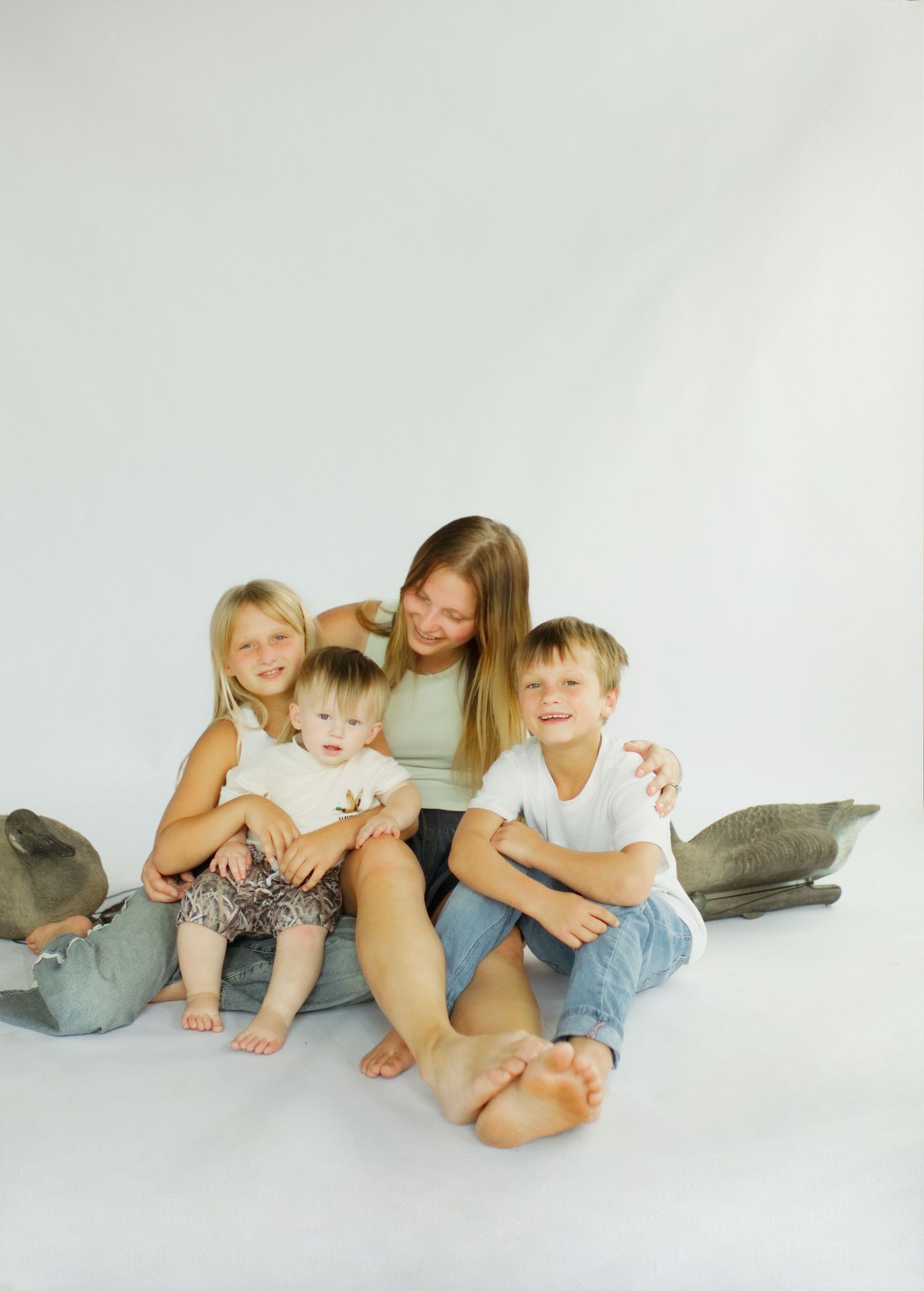 A mother sits on a white studio floor smiling with her three young children, including a toddler, in casual outfits.