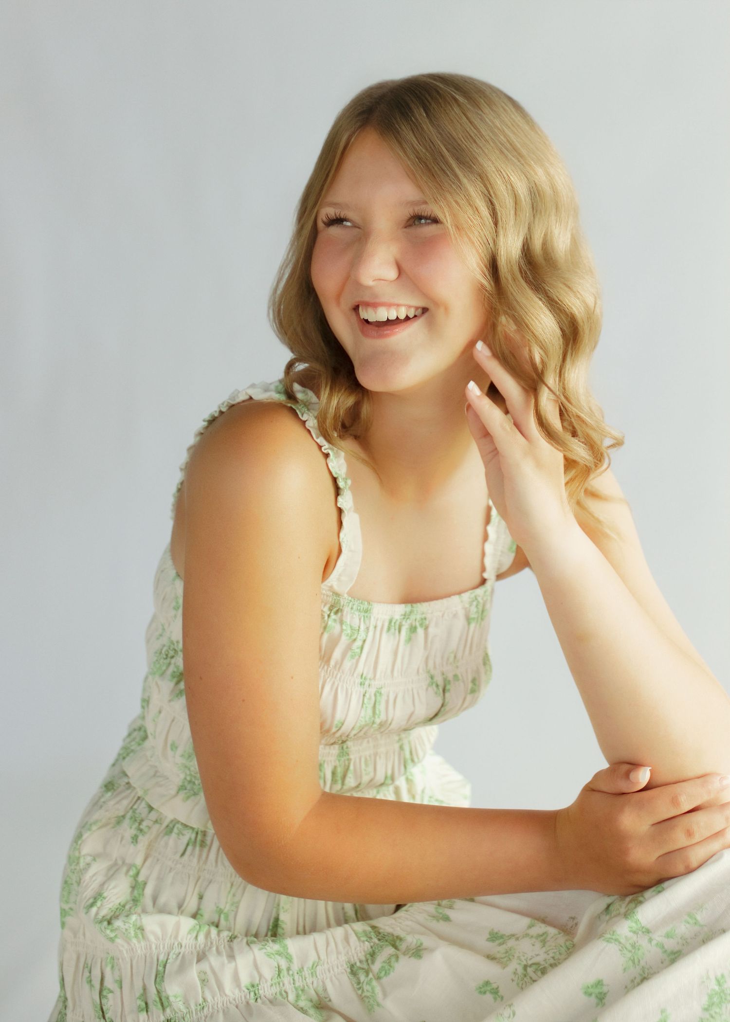 Smiling teenage girl with blonde wavy hair wearing a white floral sundress, posing against a light gray background.