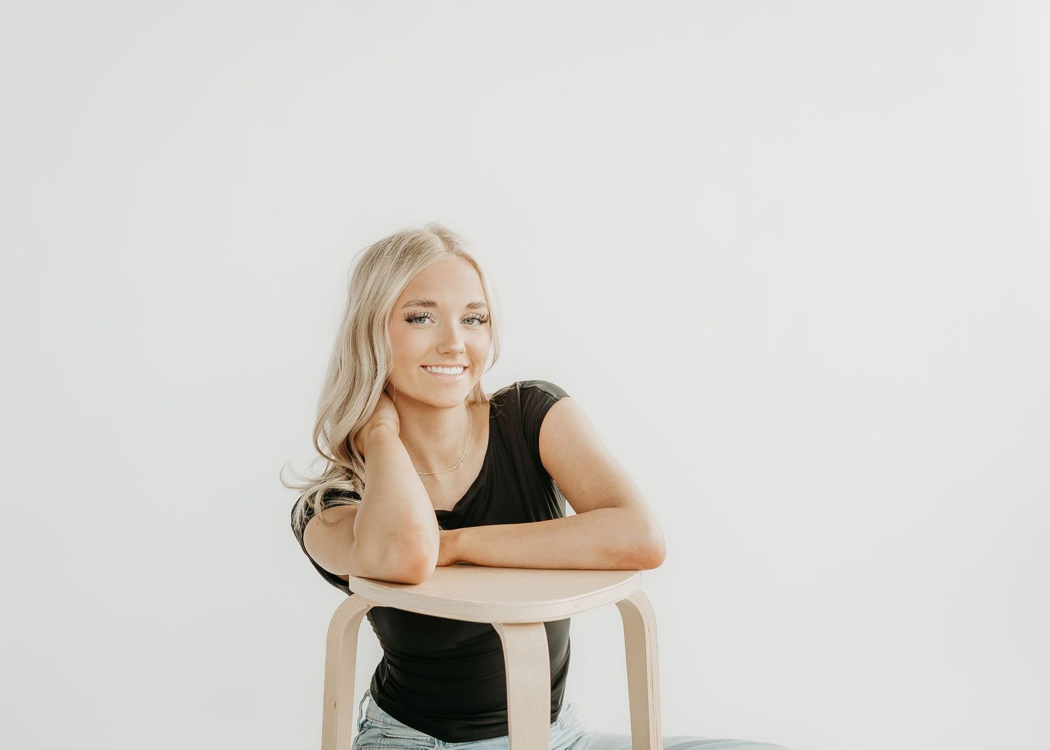 Woman in black top sits casually on wooden chair against white background in minimalist portrait series.