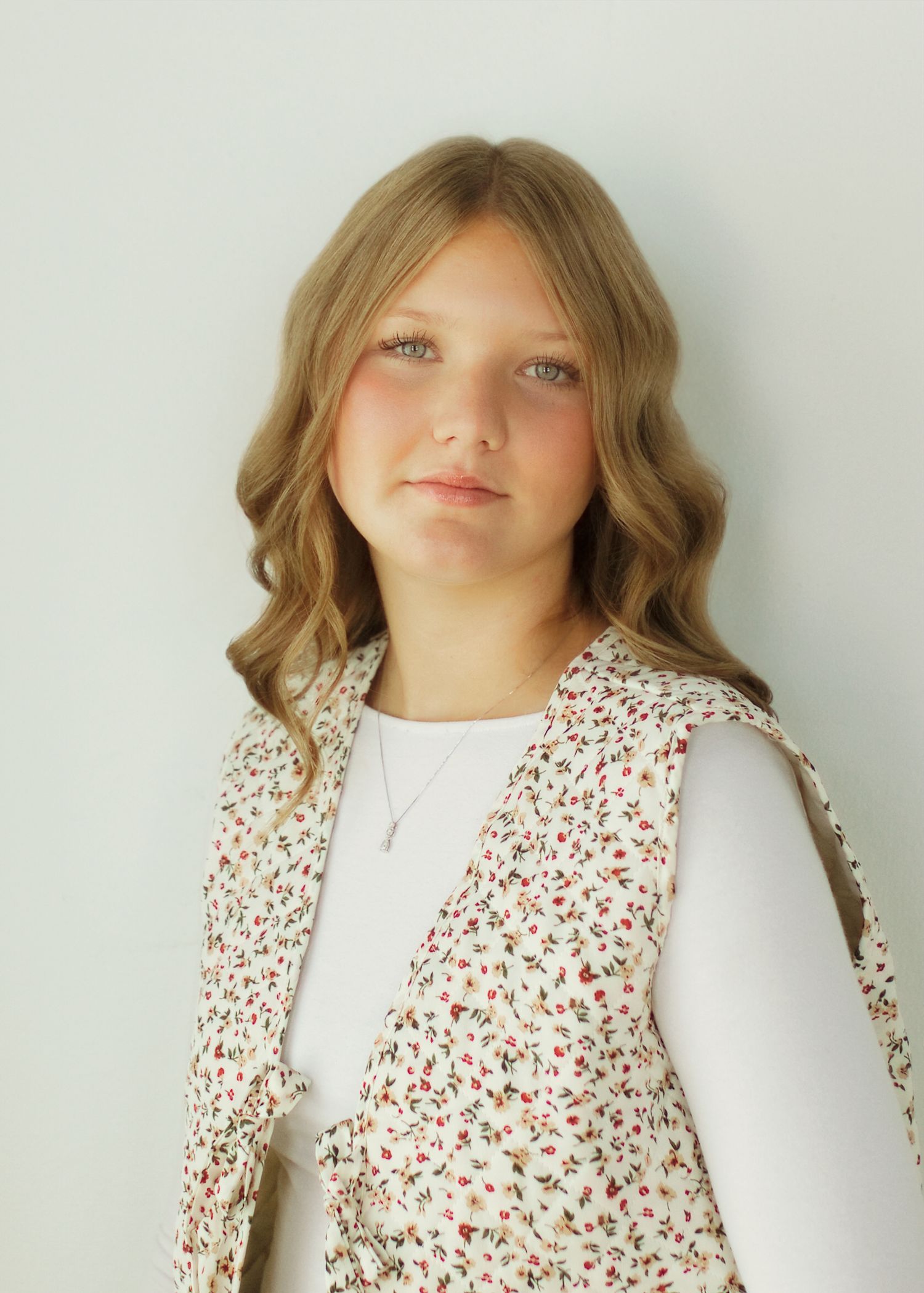Young girl with wavy blonde hair wearing a floral vest over a white top, posing for a portrait photo.