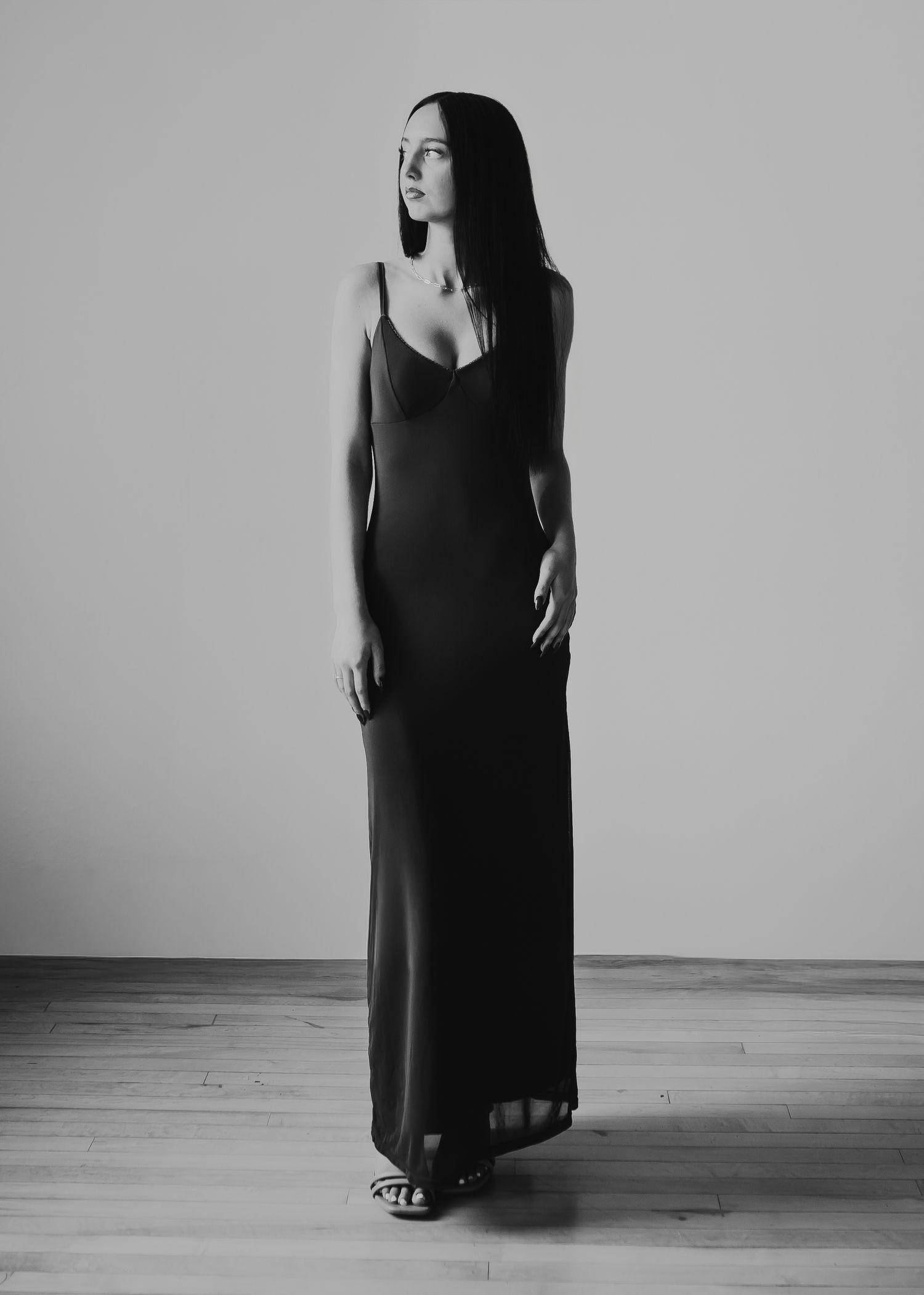 Black and white photo of a woman in a long black slip dress standing elegantly in a minimalist studio setting.