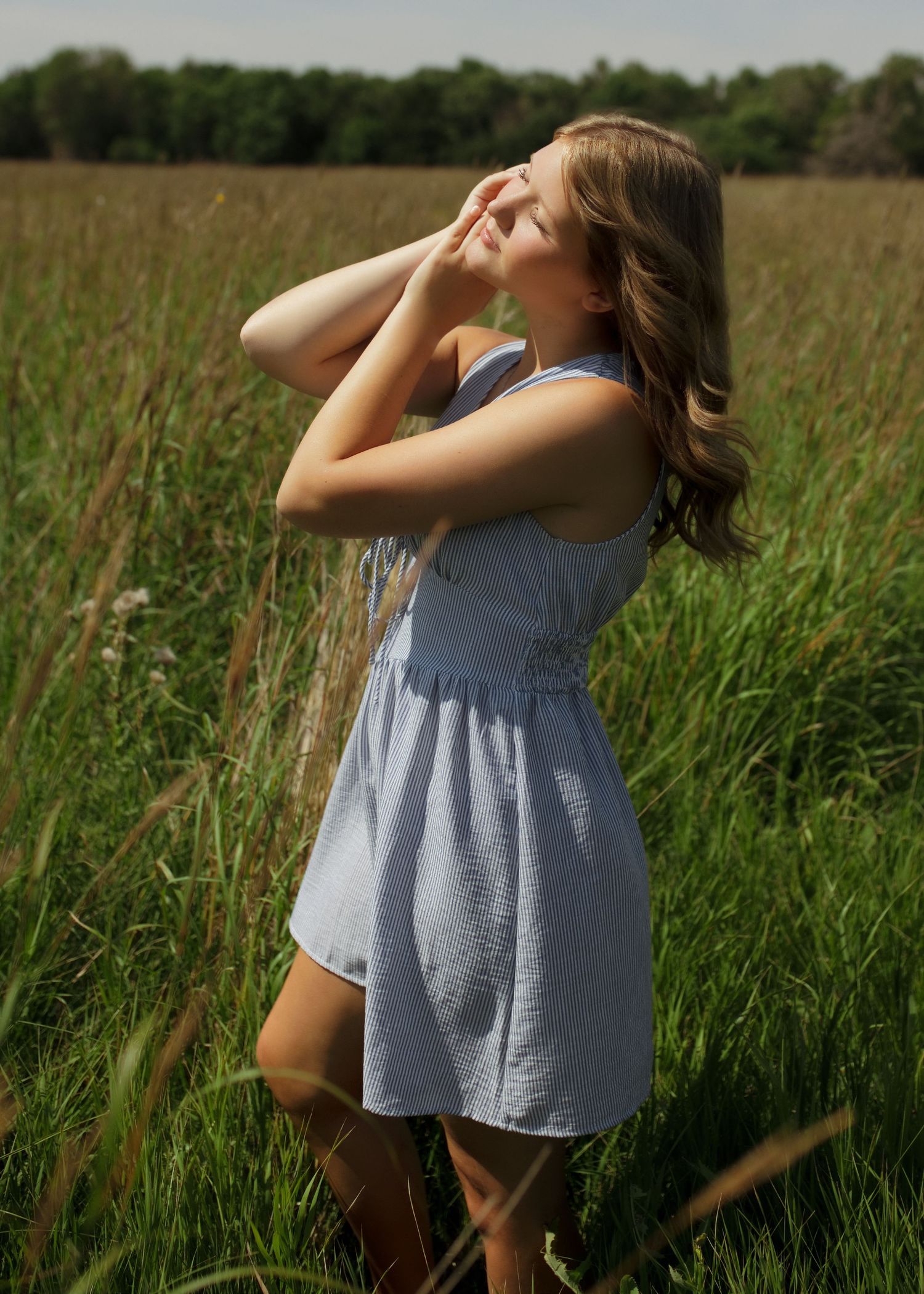 Young woman in blue dress standing in tall grass field, hands raised to face, with trees in background.