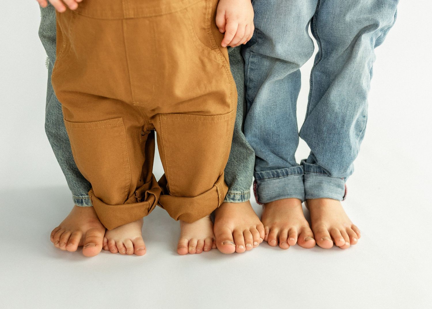 Two children kneeling side by side, one in tan overalls and one in blue jeans, showing their bare feet.